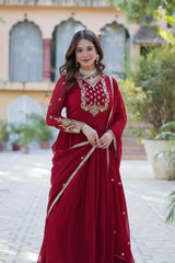 A full-length front view of a woman in a red ethnic gown with gold embroidery. She is looking toward the camera with a gentle smile, wearing a pearl and gold choker necklace. The background shows a building with traditional arched windows and greenery.
