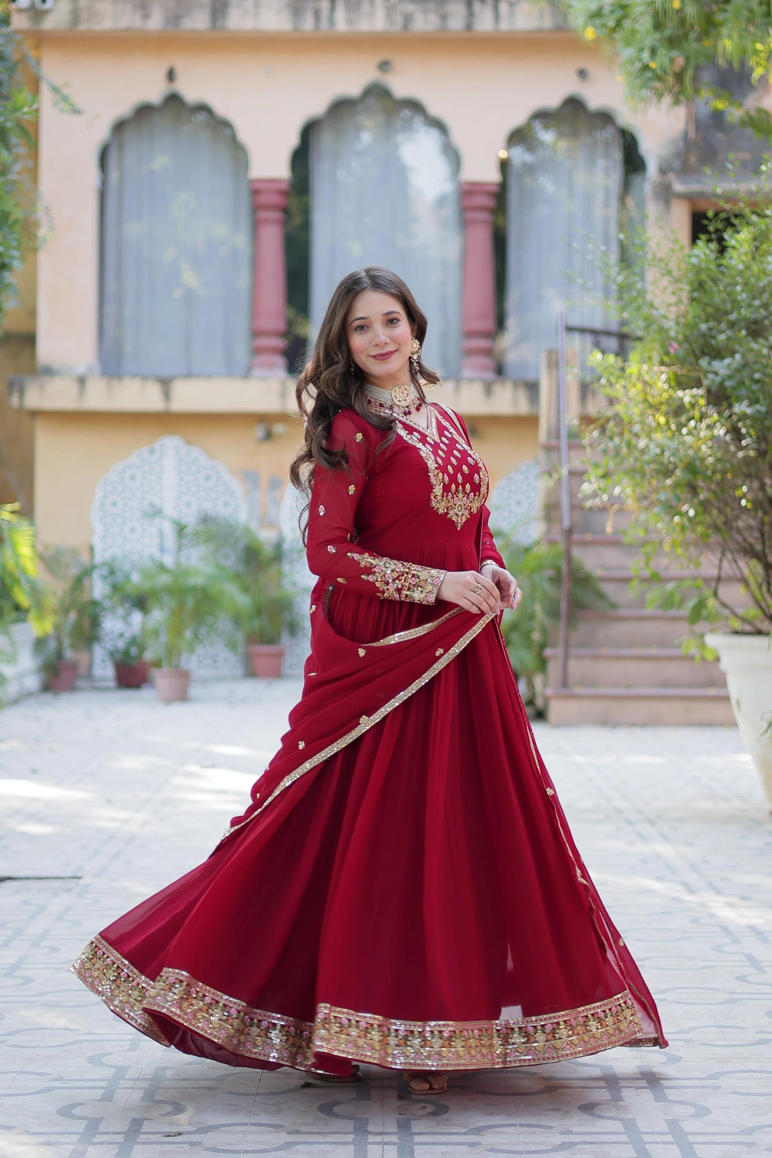 A woman stands in a tiled courtyard wearing a deep red floor-length Anarkali suit. The dress features gold floral embroidery on the bodice, sleeves, and a heavy decorative border at the hem. She is draped in a matching red dupatta with gold piping.