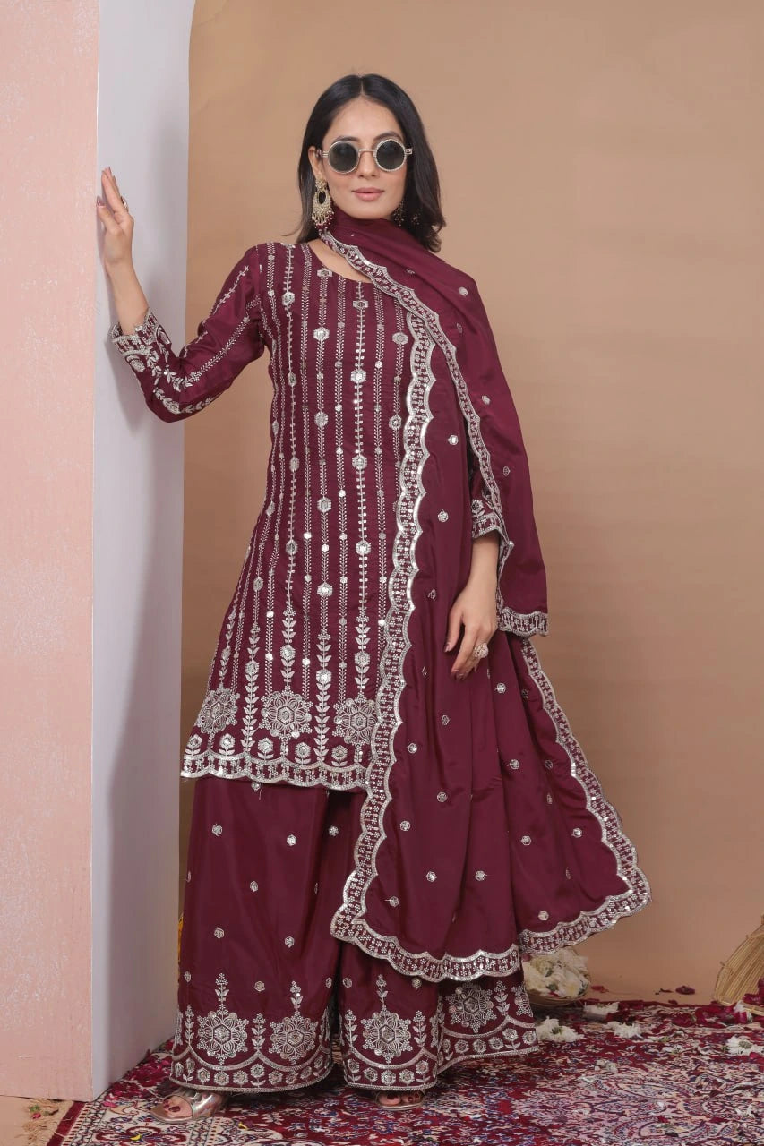 Woman wearing a maroon traditional outfit with white patterns against a beige wall.