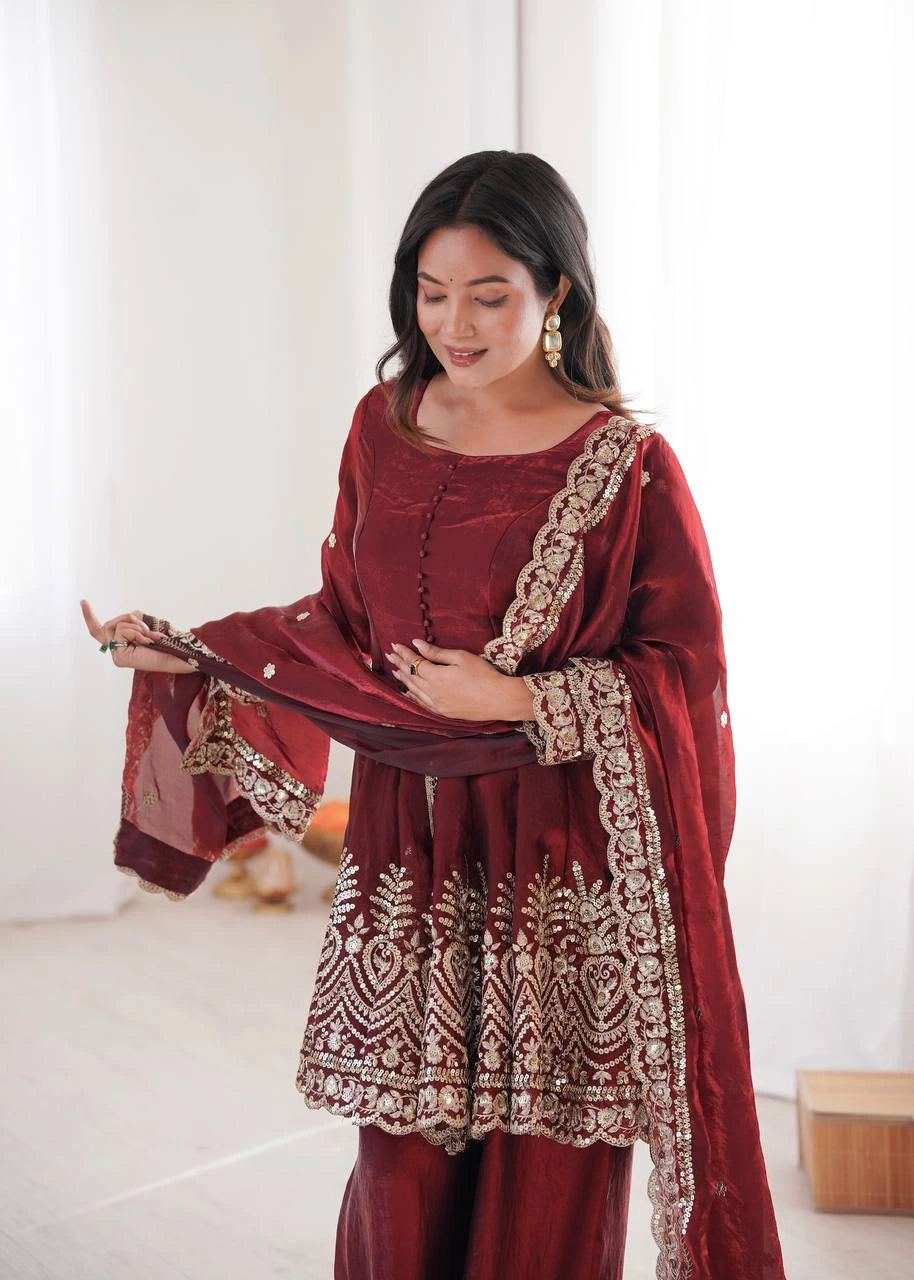 Woman wearing a maroon traditional outfit with gold embroidery in a minimal indoor setting.