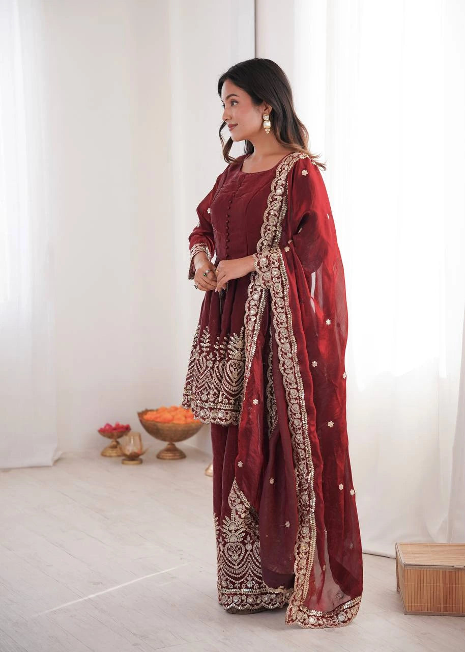 Woman in a traditional maroon outfit with gold embroidery standing in a minimalistic room.