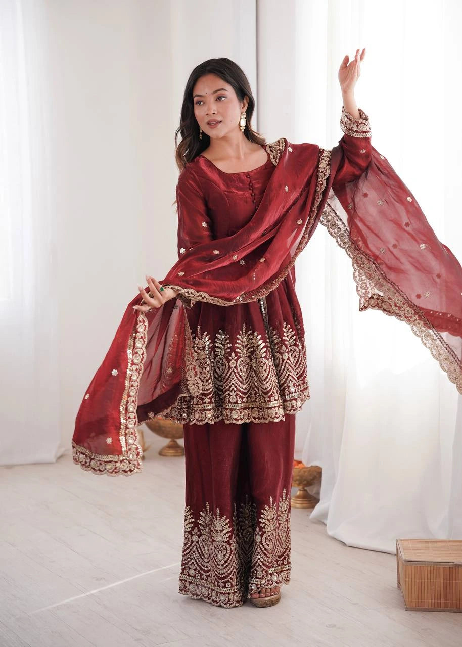 Woman wearing a traditional maroon outfit with gold embroidery in a minimalistic setting.