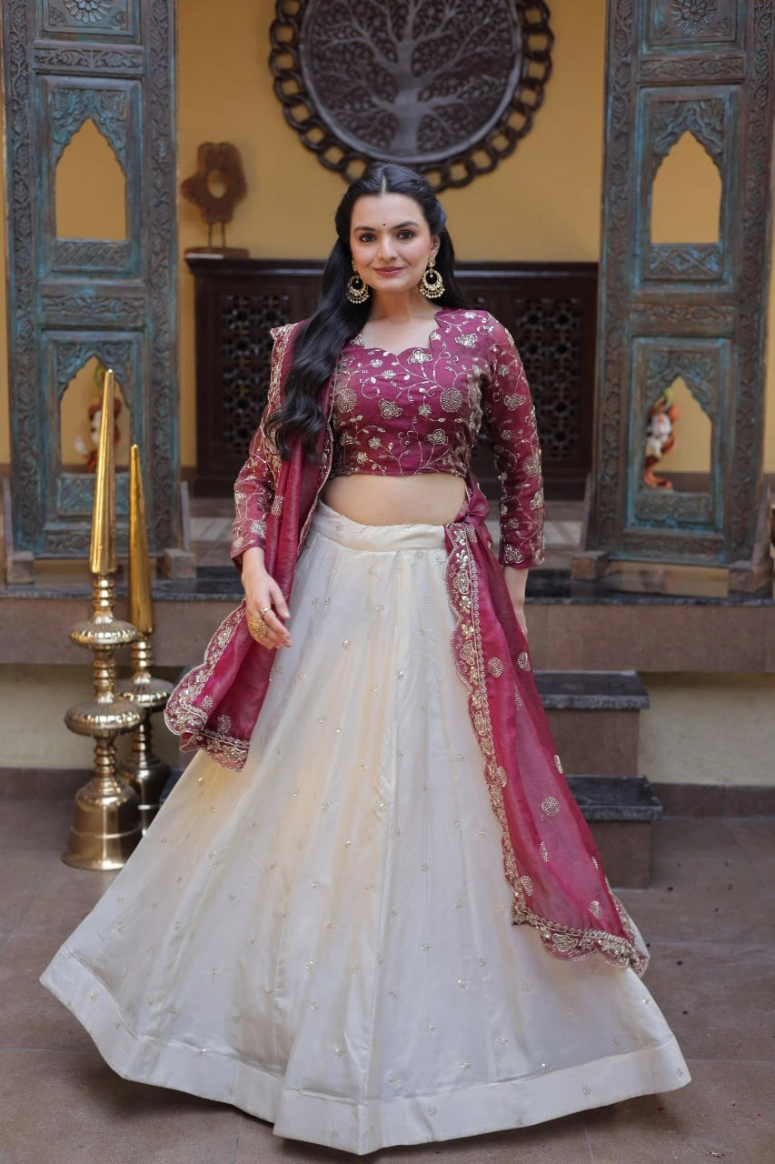 Woman in a traditional outfit with pink blouse and white lehenga standing in a decorative interior setting.