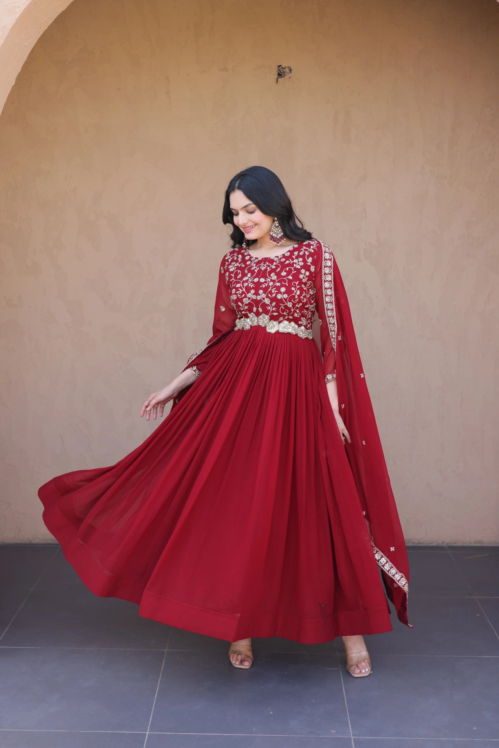 Woman wearing a red traditional outfit with gold embroidery against a beige wall.