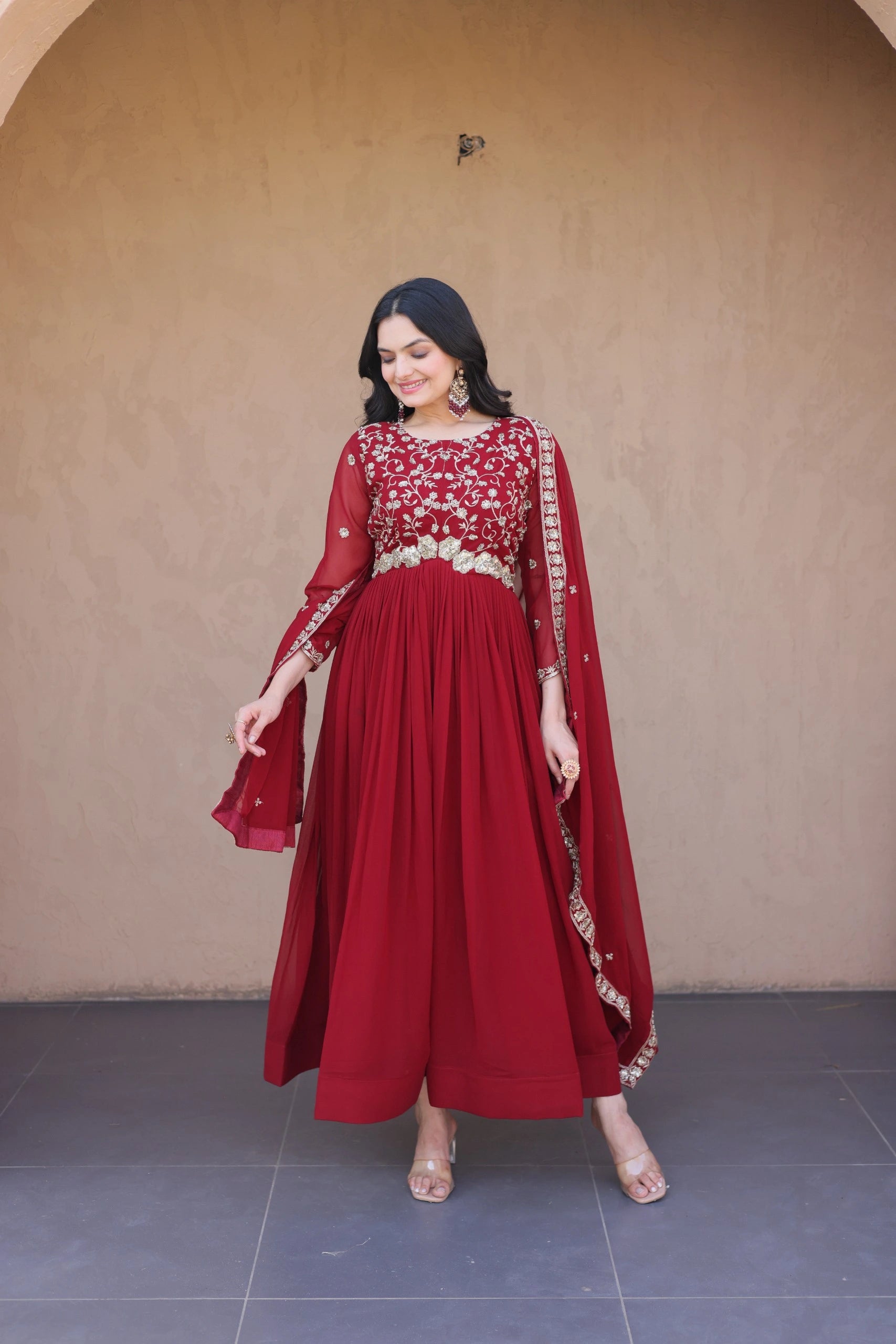 Woman wearing a red traditional outfit with gold details against a beige wall.