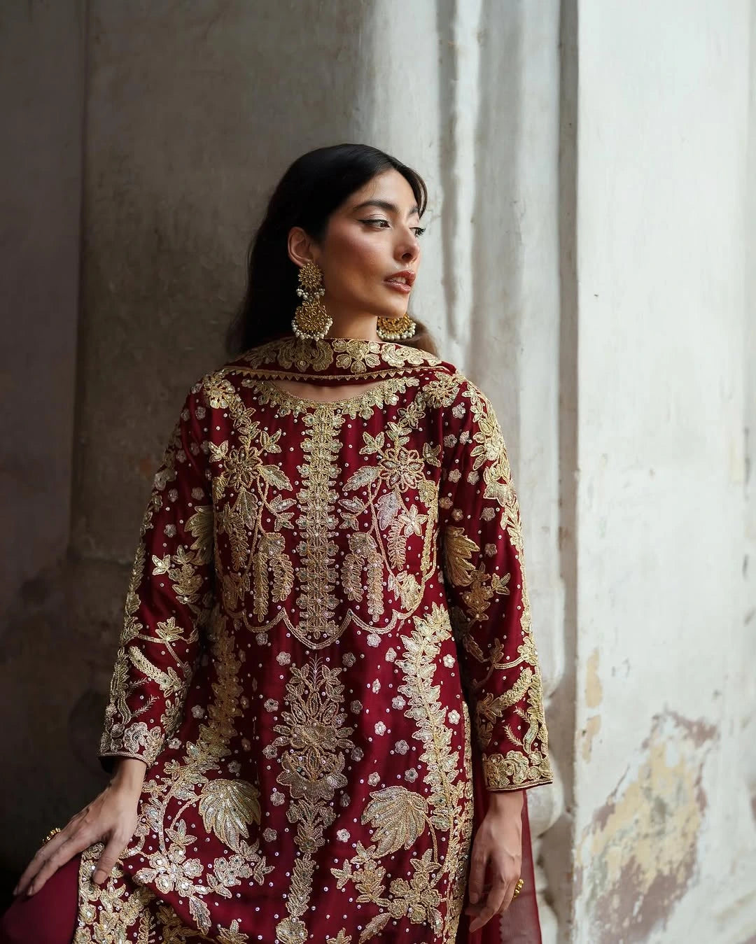 Woman wearing a red and gold embroidered traditional outfit against a textured wall.