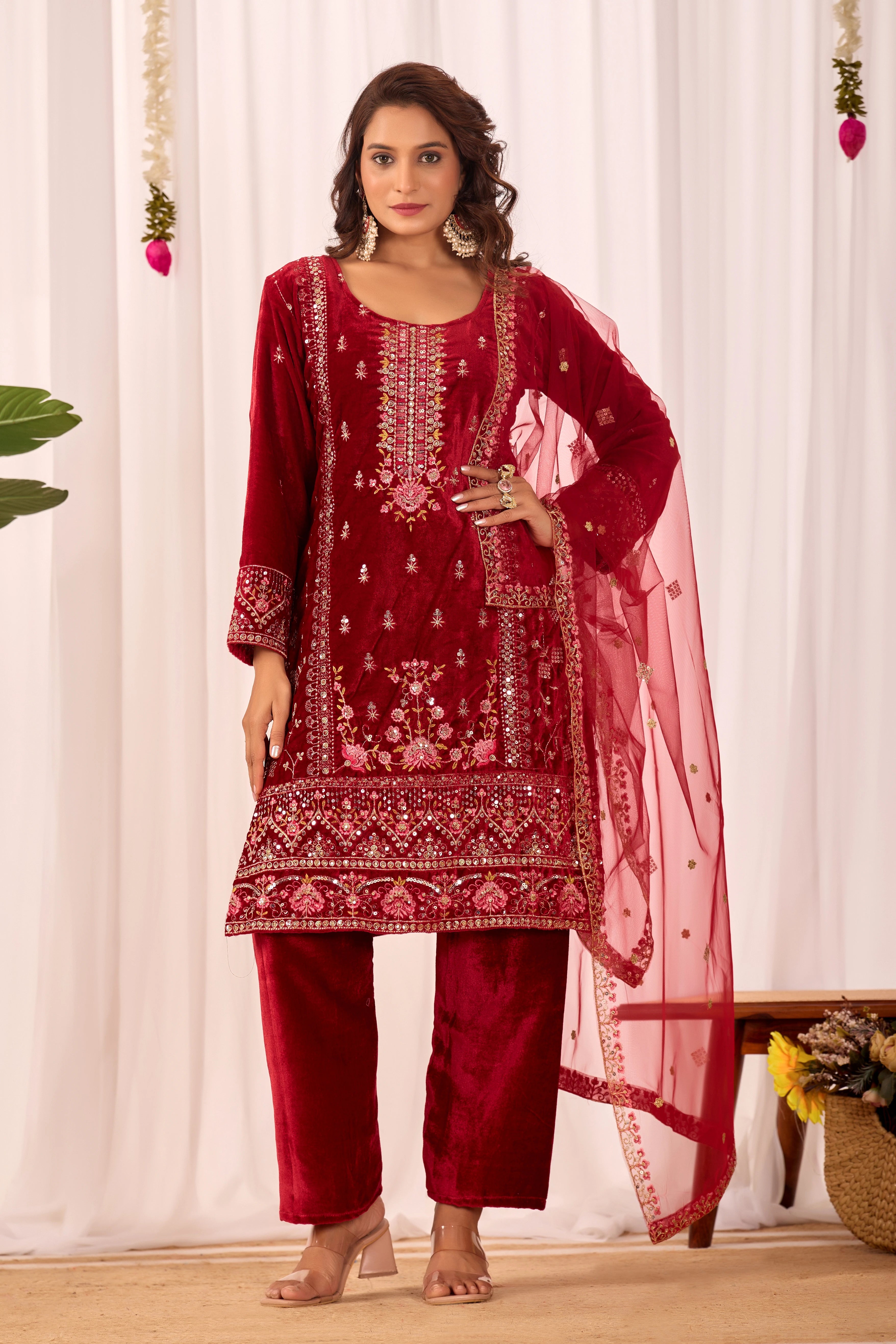 Woman wearing a red embroidered traditional outfit with a dupatta against a white curtain background