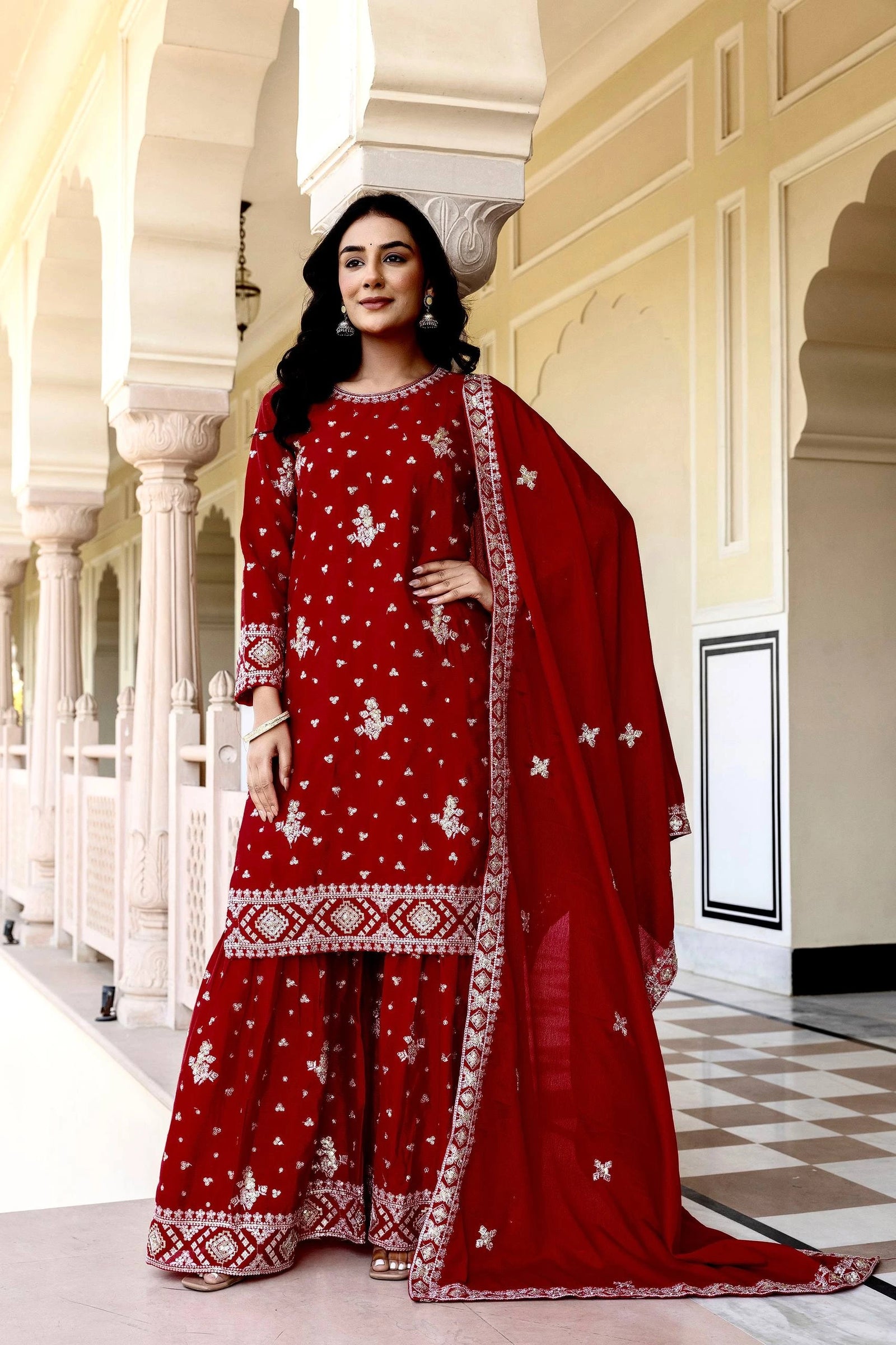Woman in a red traditional outfit with white patterns standing in an elegant hallway.