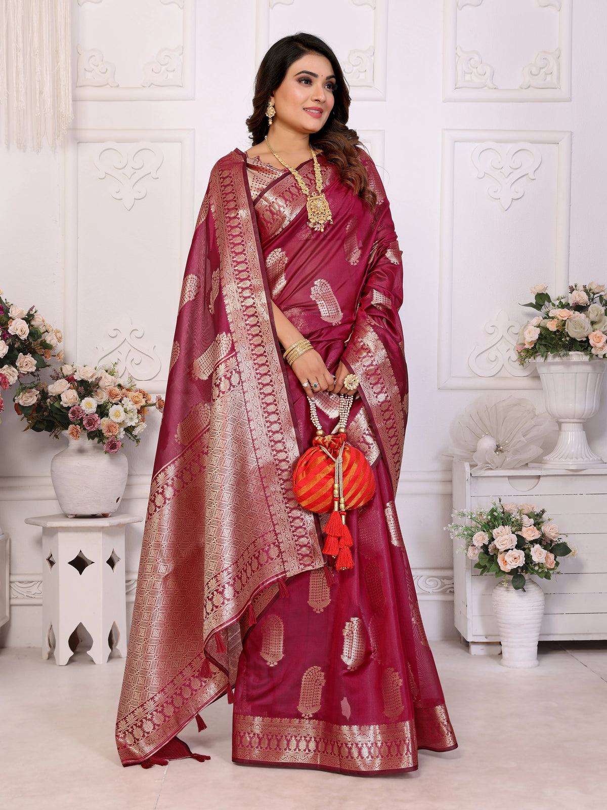 Woman wearing a red saree with gold patterns in a decorated room.