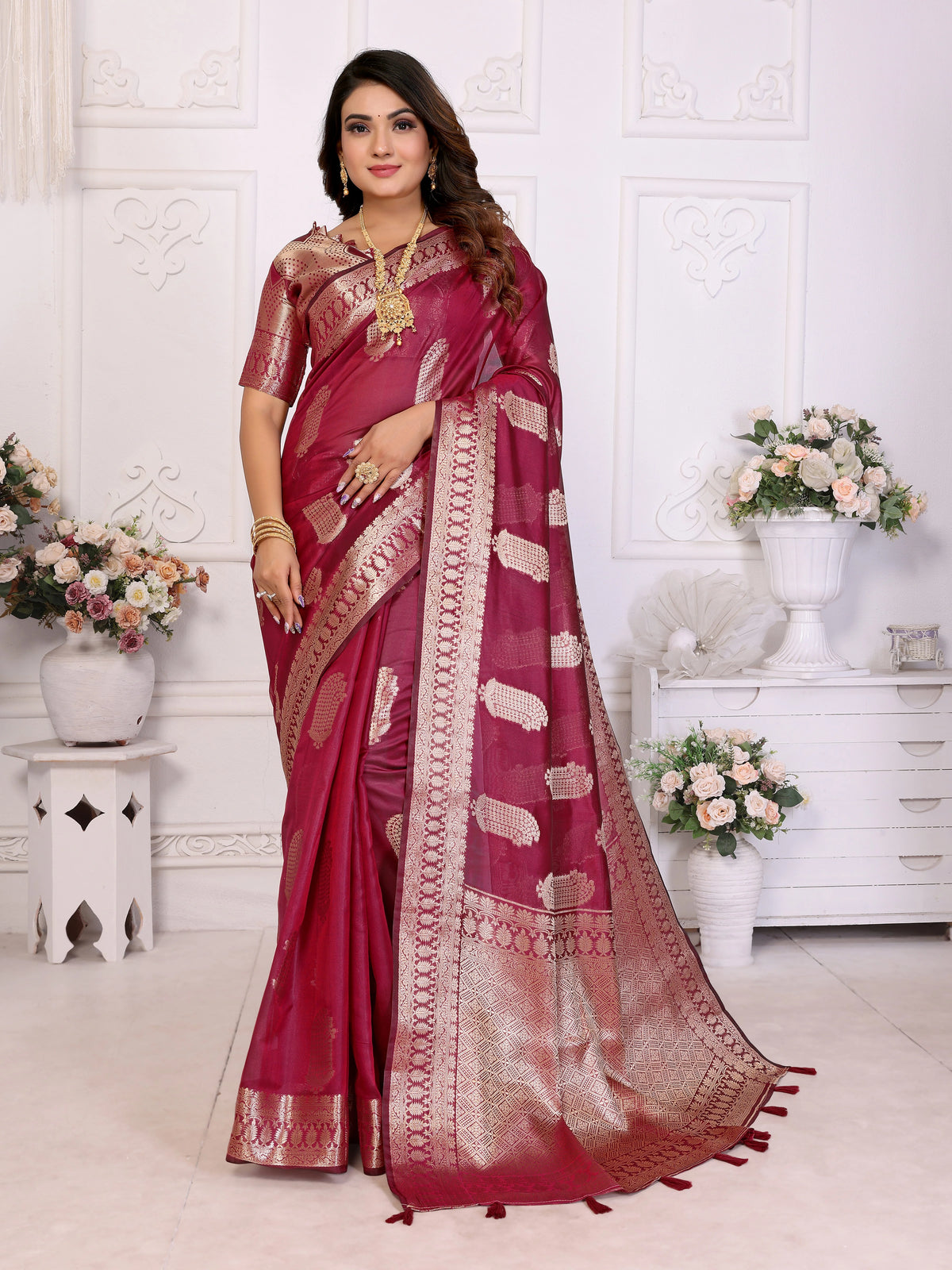 Woman wearing a red saree with gold patterns in a decorated room.