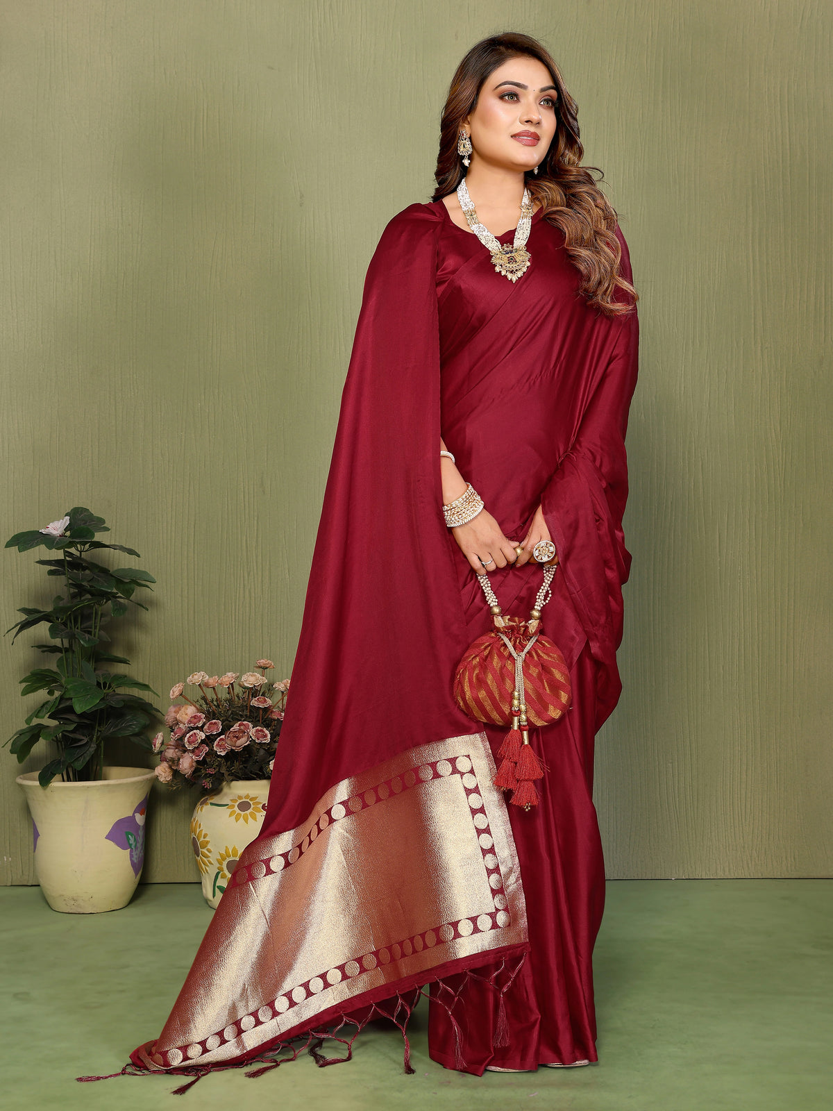 Woman in a red saree with gold border holding a matching handbag against a green background