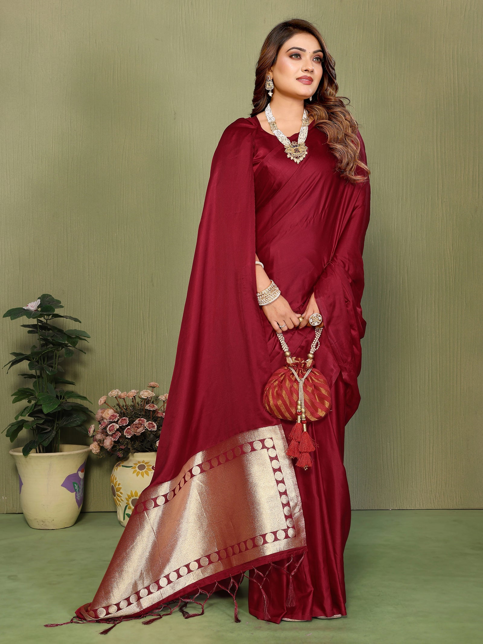 Woman in a red saree with gold border holding a matching handbag against a green background