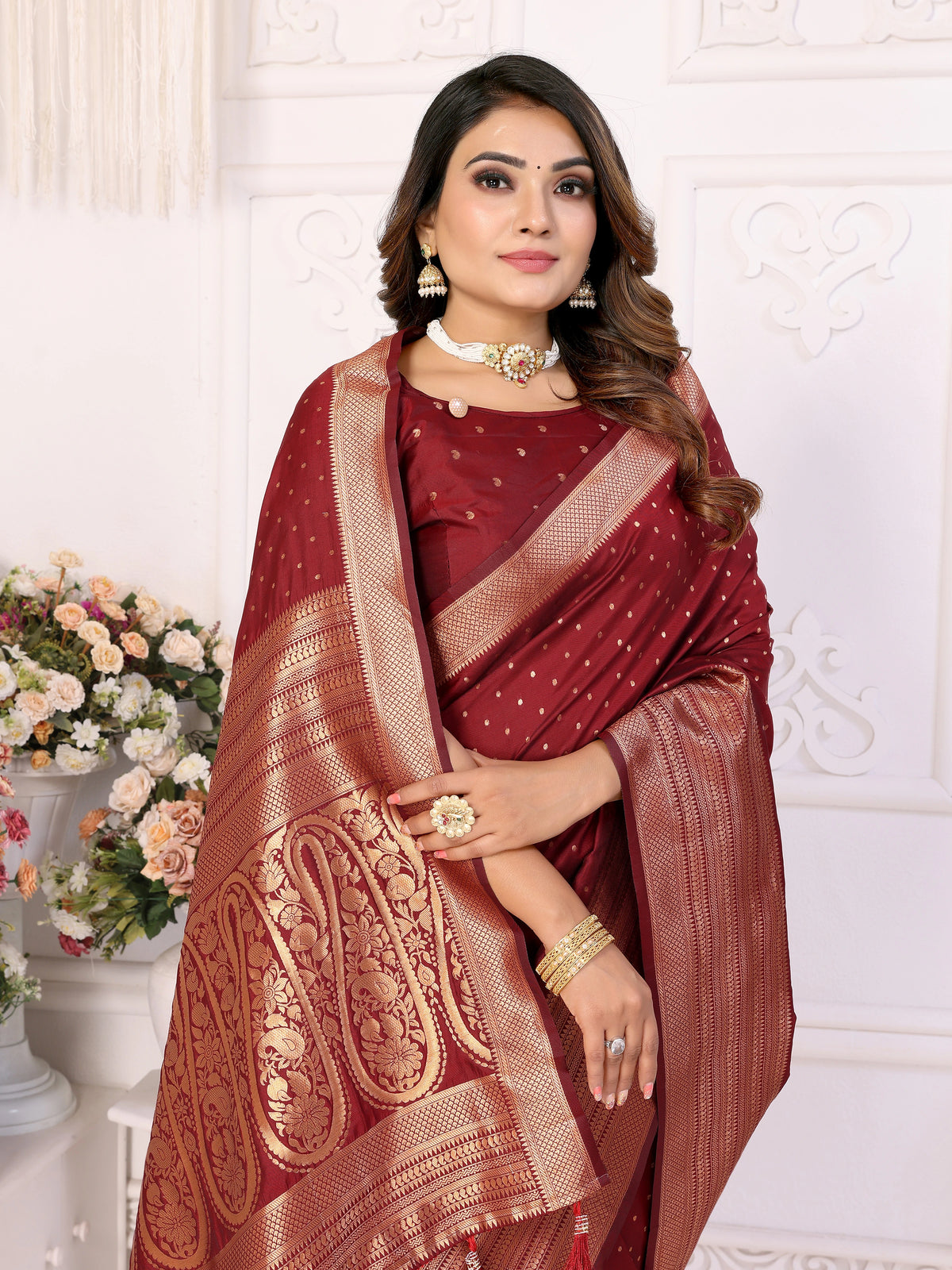 Woman wearing a maroon saree with gold patterns against a white decorative wall.