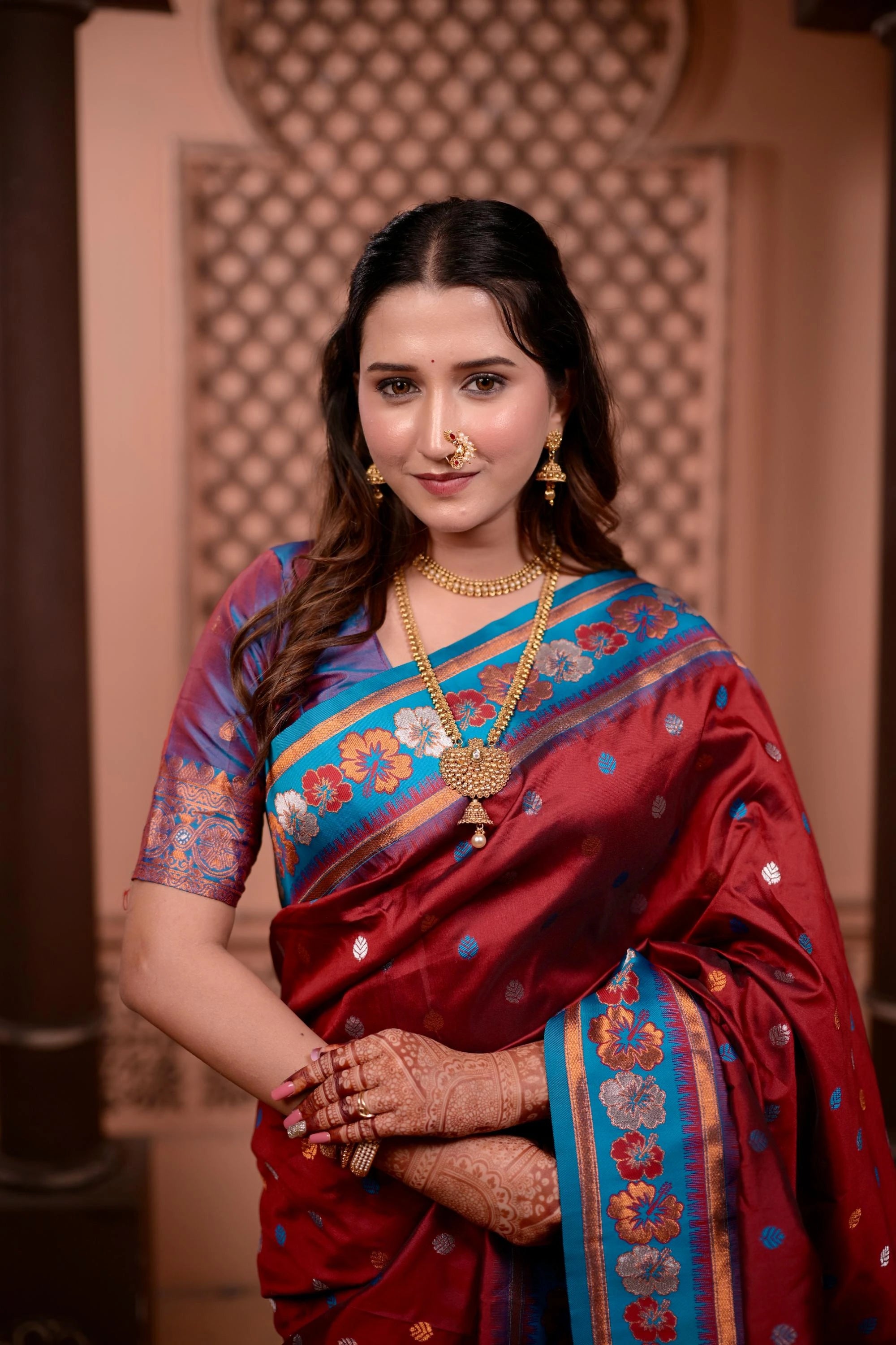 Woman in a traditional saree with intricate patterns and jewelry against an ornate wall.