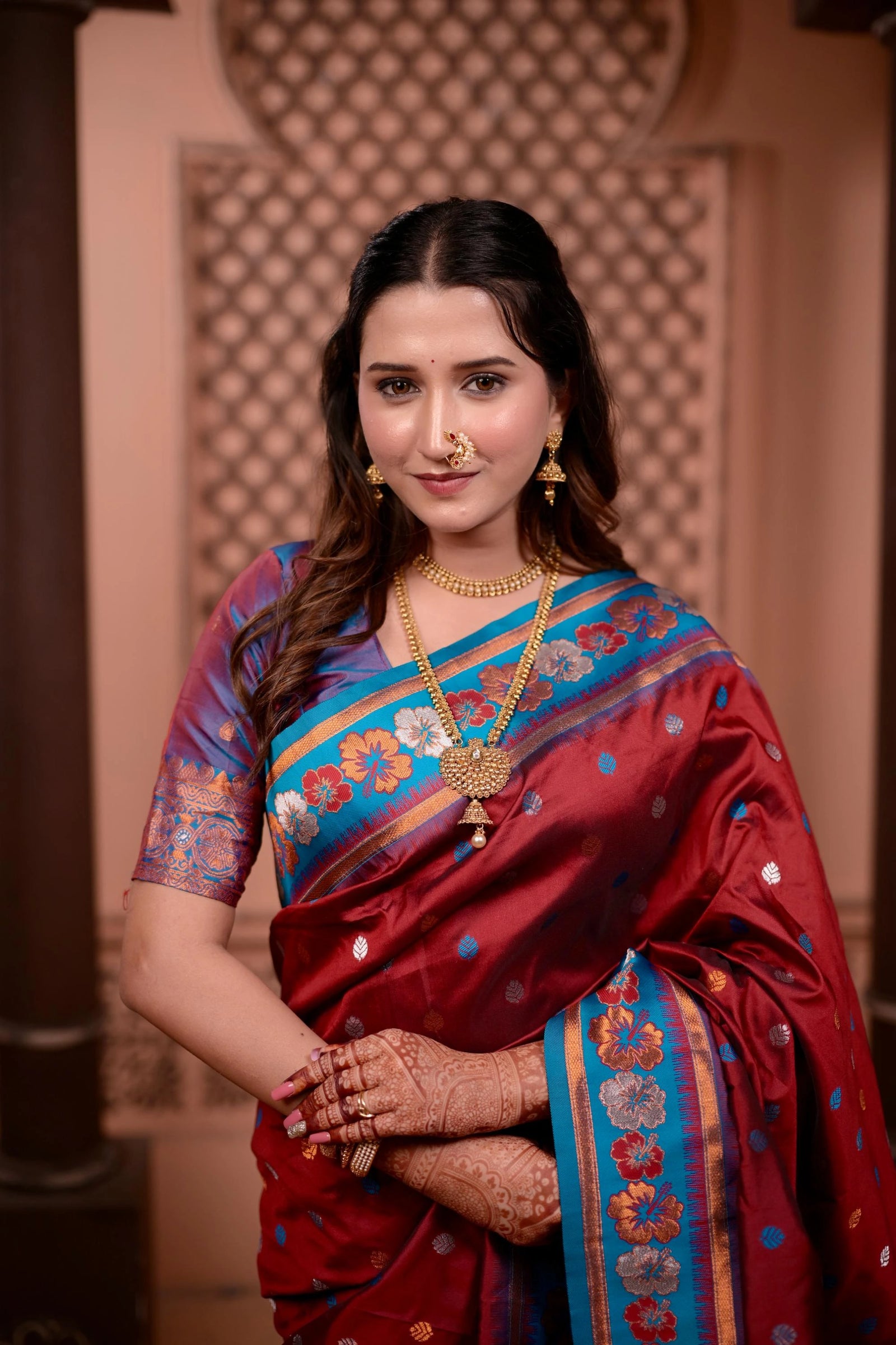 Woman in a traditional saree with intricate patterns and jewelry against an ornate wall.