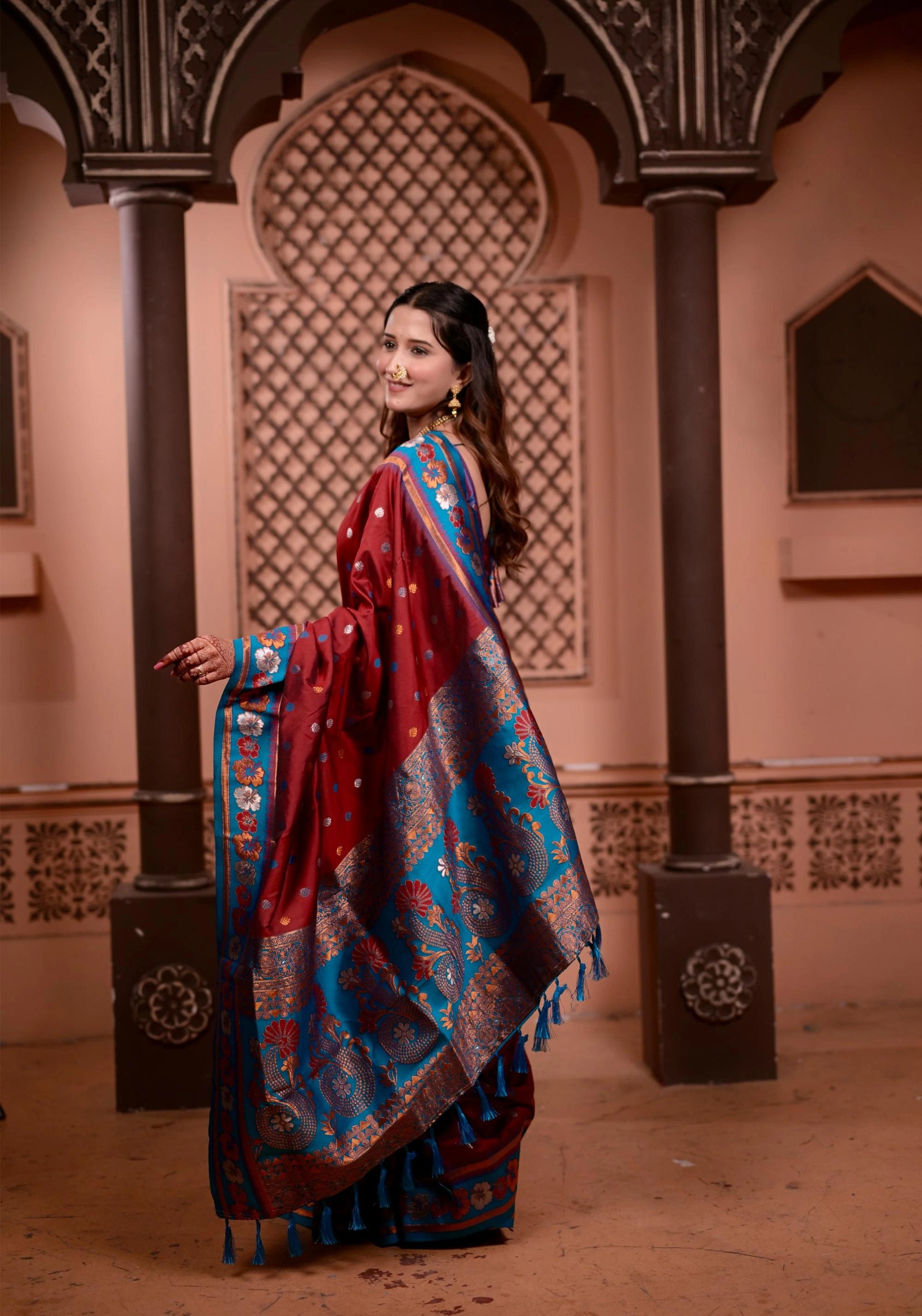 Woman in a red and blue saree standing in an ornate indoor setting.