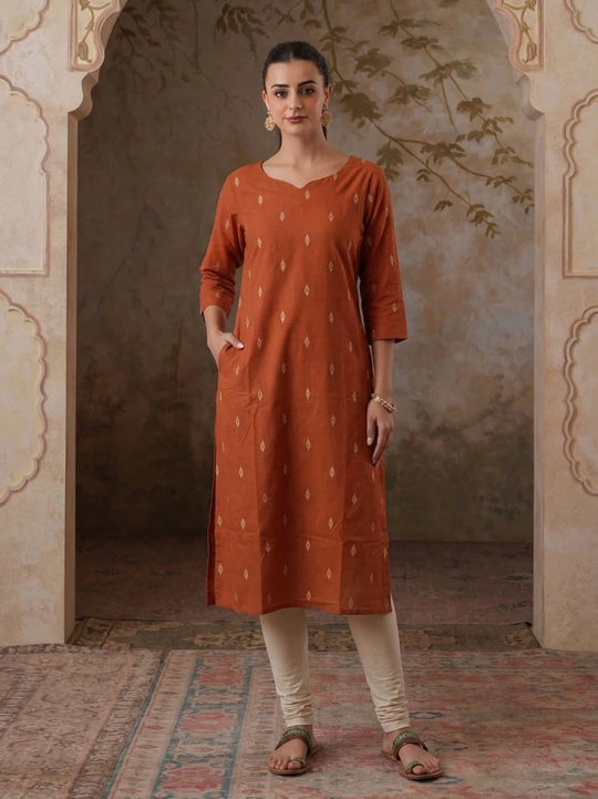 Woman wearing a rust-colored kurta with white patterns in an ornate room.