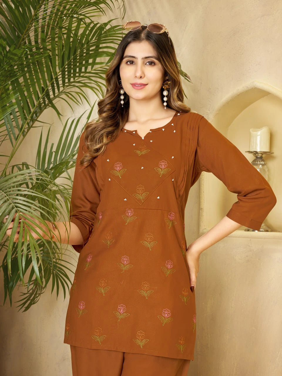 Woman wearing a brown embroidered outfit standing next to a plant indoors.