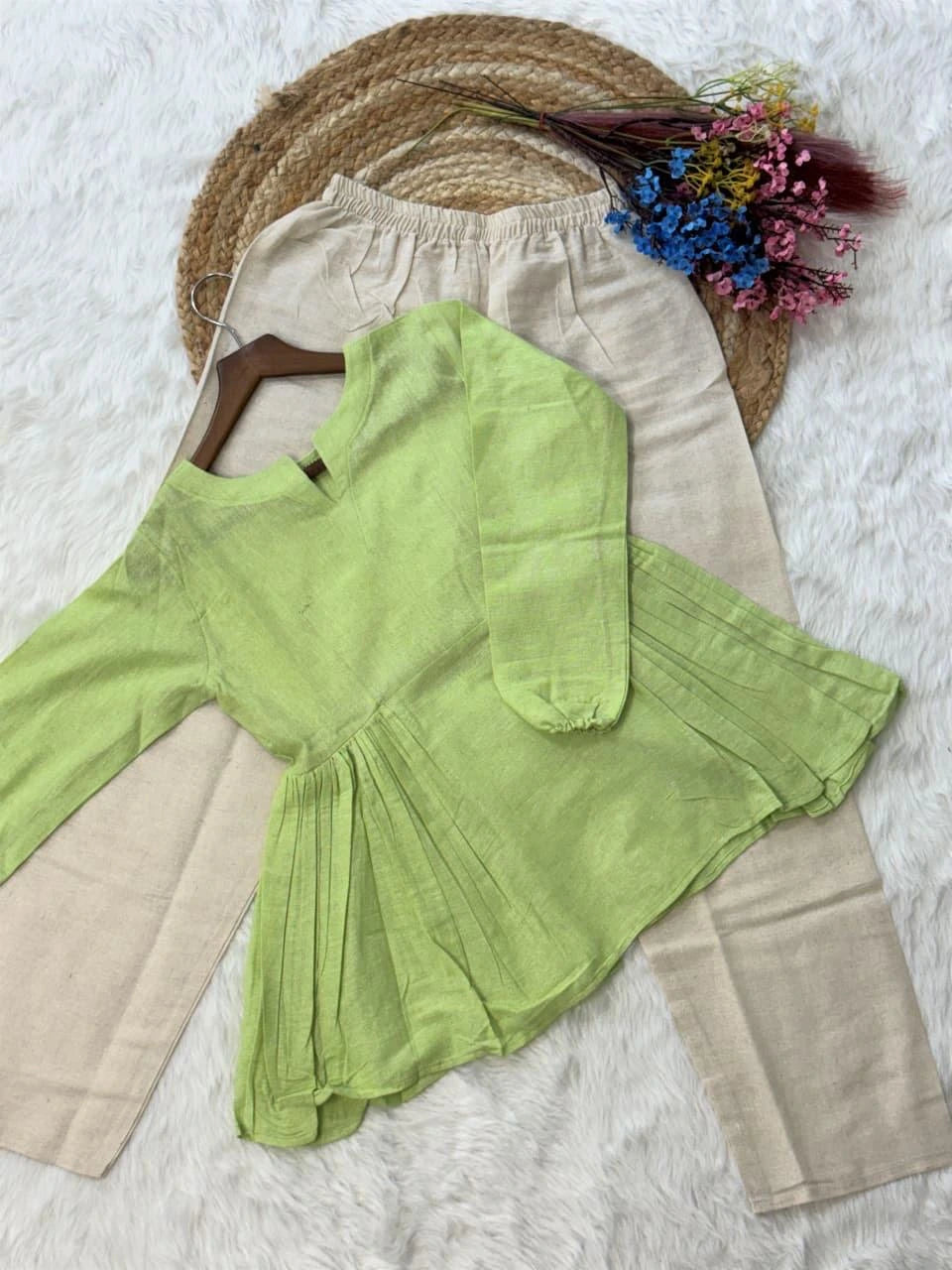 Green top and beige pants on a white surface with a straw hat and flowers.