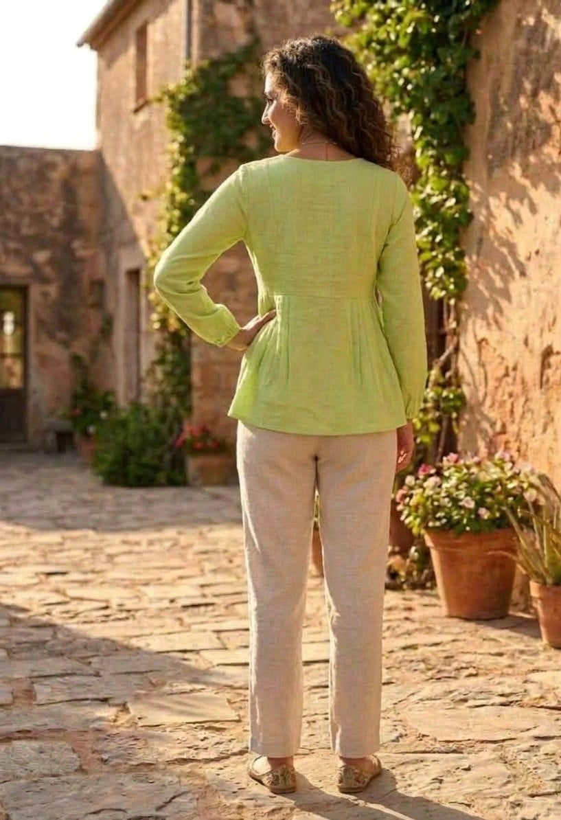 Woman in a green top and beige pants standing in a sunlit outdoor setting with stone walls and plants.