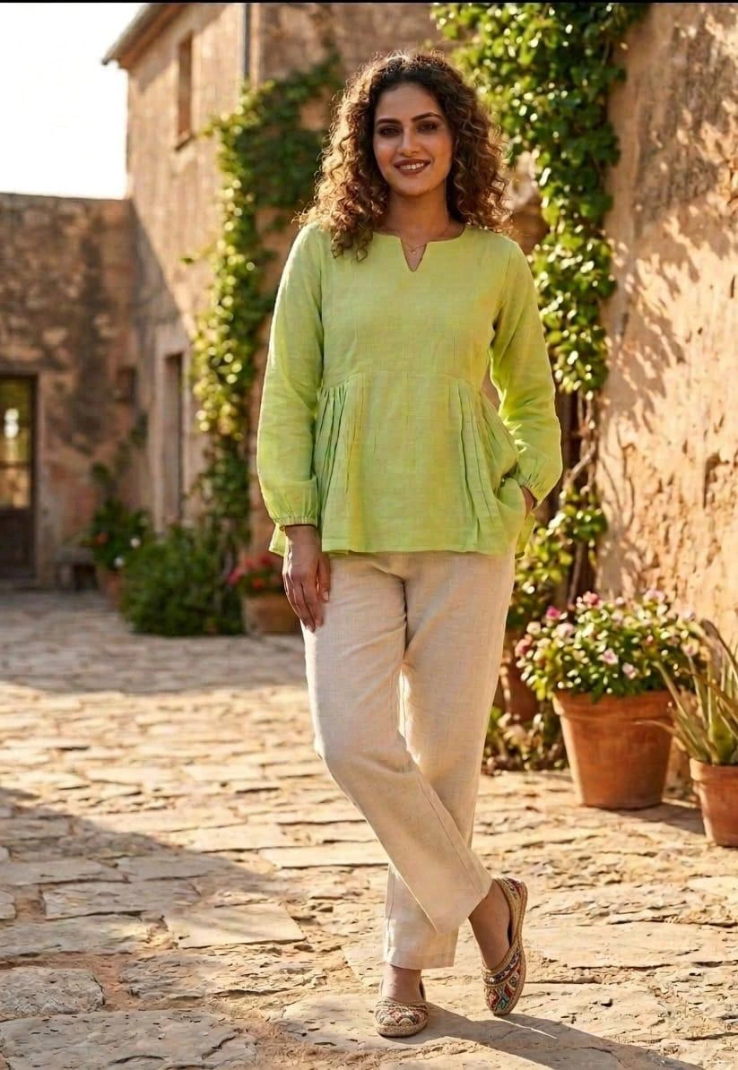 Woman in a light green top and beige pants standing outdoors with stone pavement and plants.