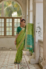 Woman in a green saree standing in an ornate room with large windows.