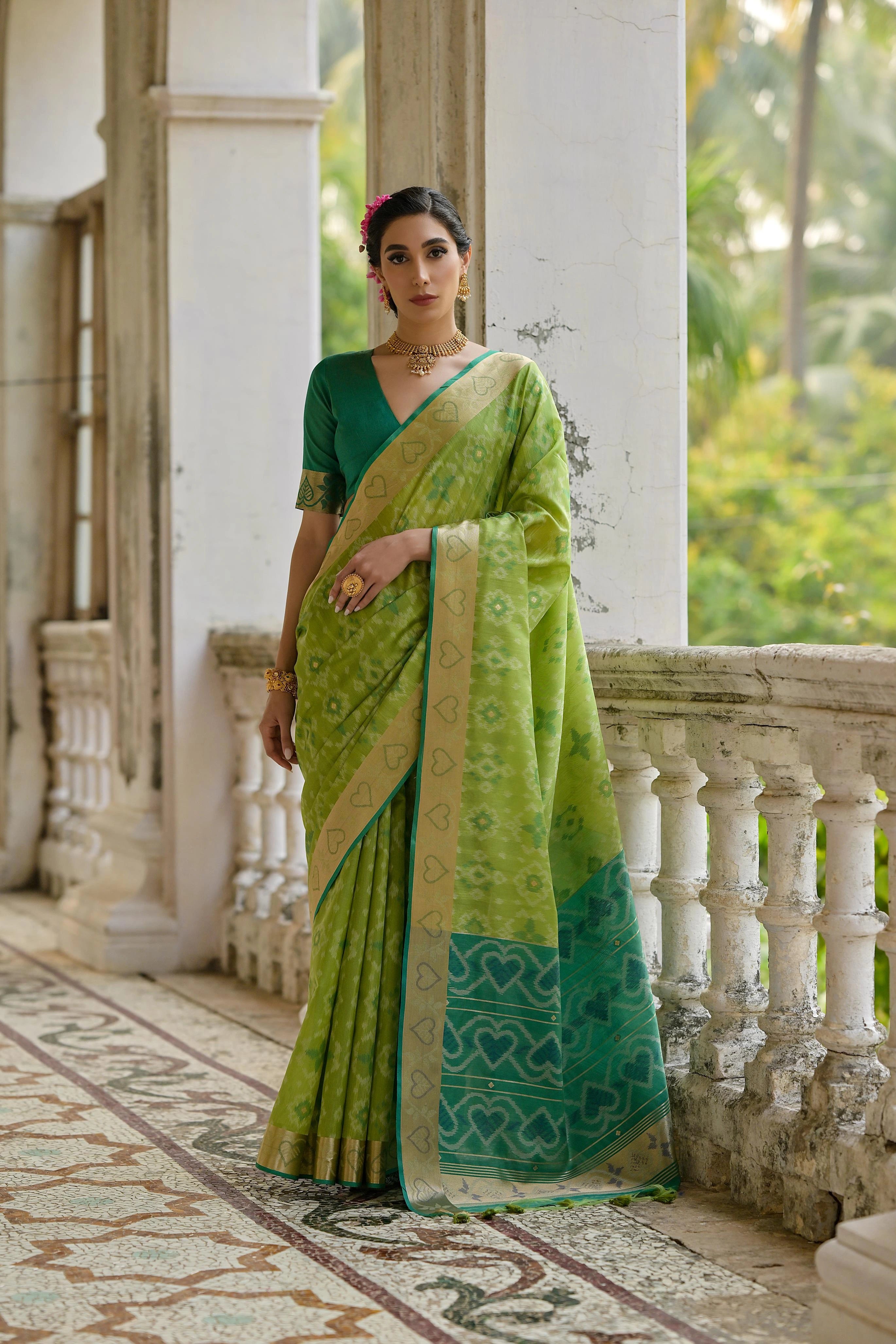 Woman in a green saree standing on a balcony with a white wall and greenery in the background