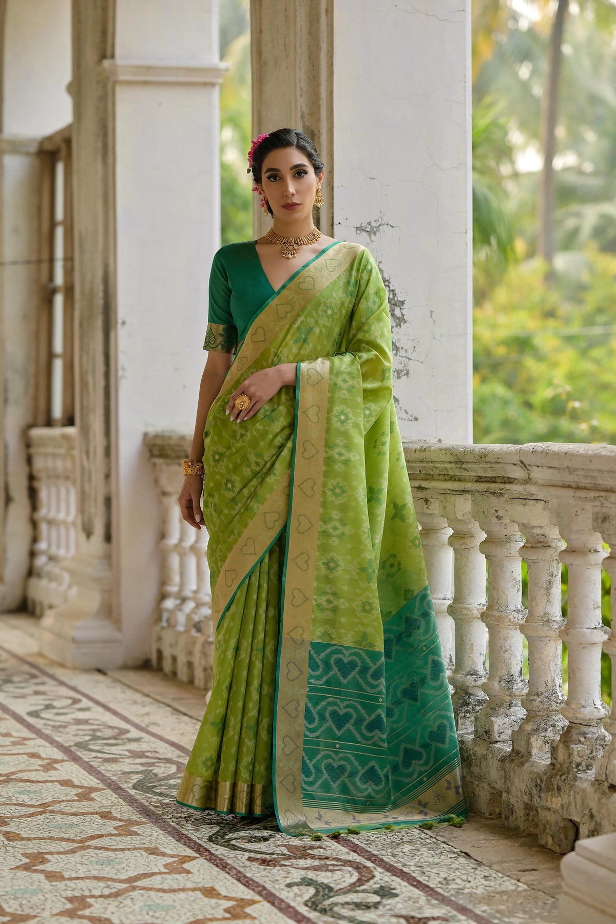 Woman in a green saree standing on a balcony with a white wall and greenery in the background