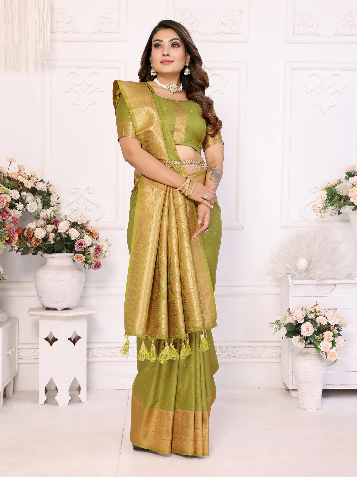 Woman in a green saree standing in a decorated room with flowers and white furniture.