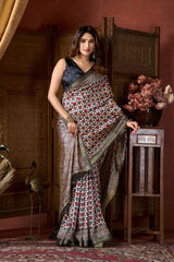 Woman in a patterned saree standing in an elegant room with wooden furniture and decorative elements.