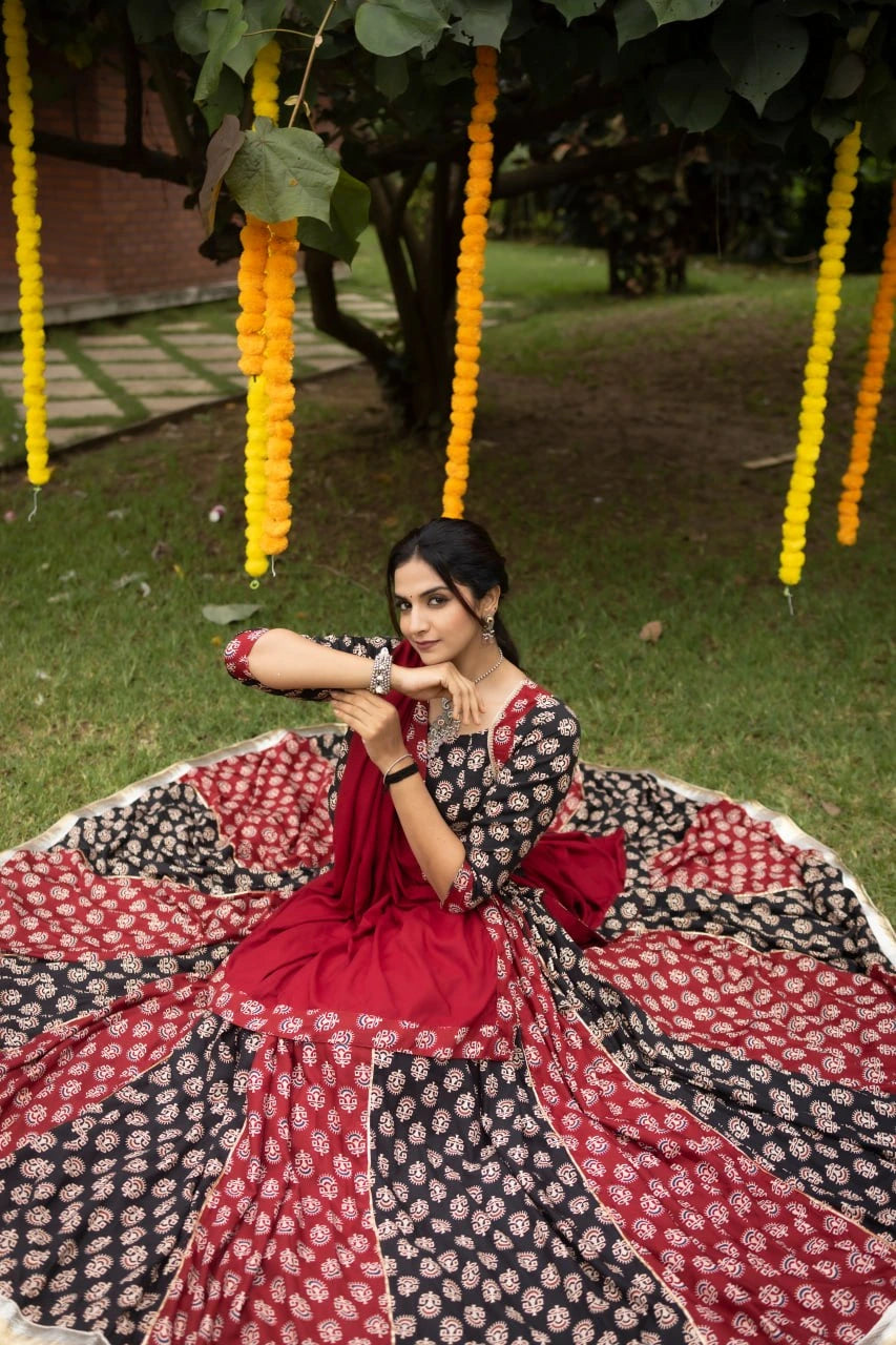 Woman in a red and black patterned saree sitting on a grassy area with decorative yellow flowers hanging above.