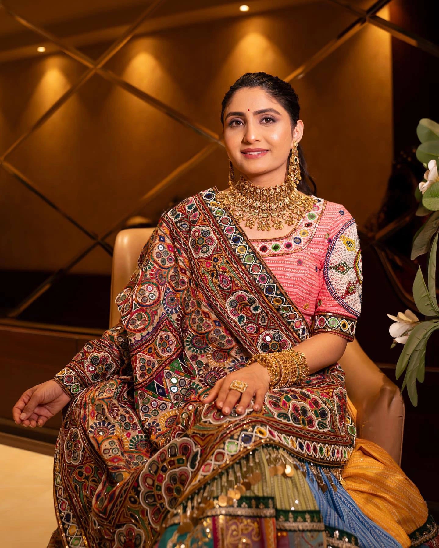 Woman in traditional attire with intricate patterns and jewelry, sitting indoors.
