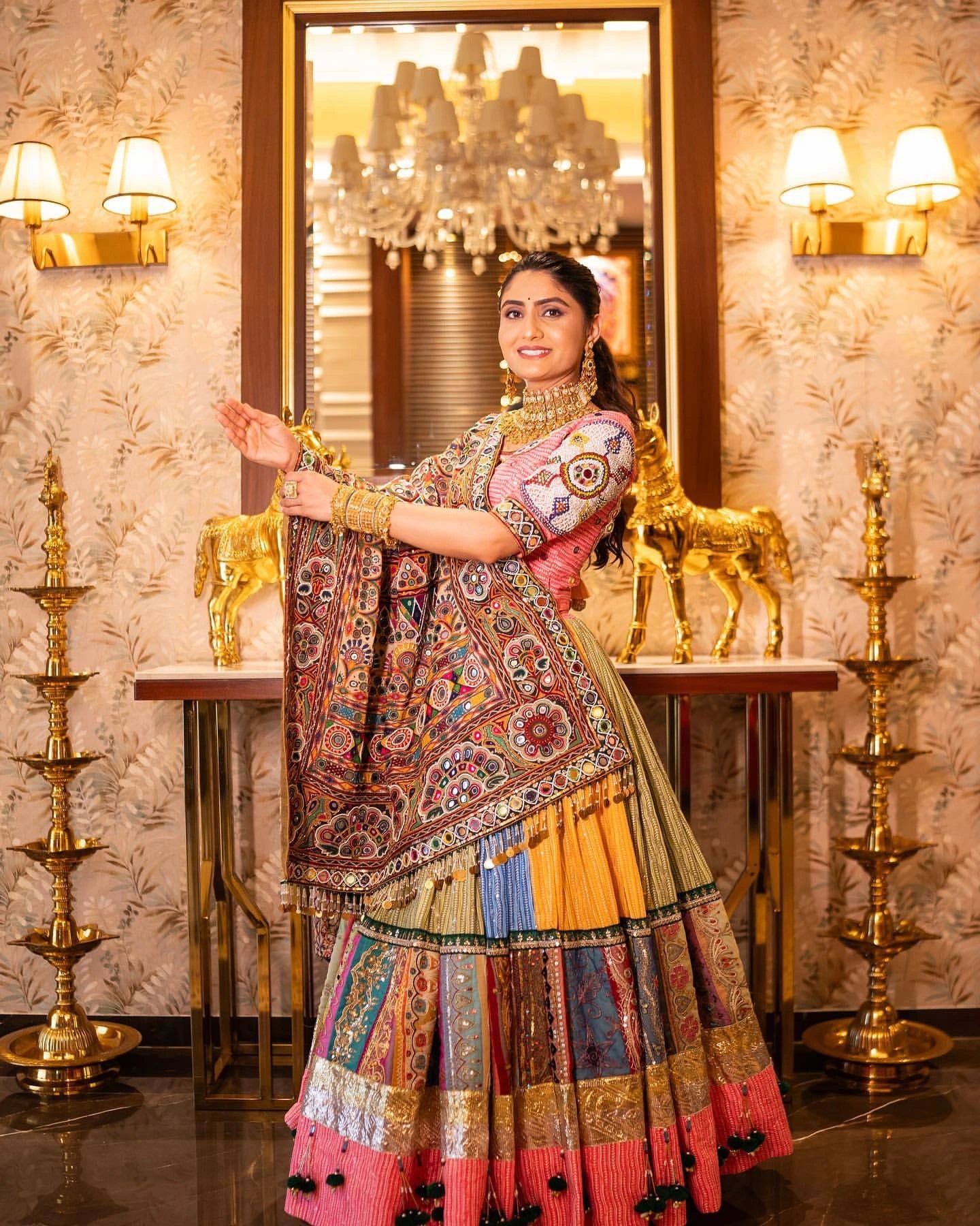 Woman in a colorful traditional outfit standing in a decorated room with chandeliers and wall lights.