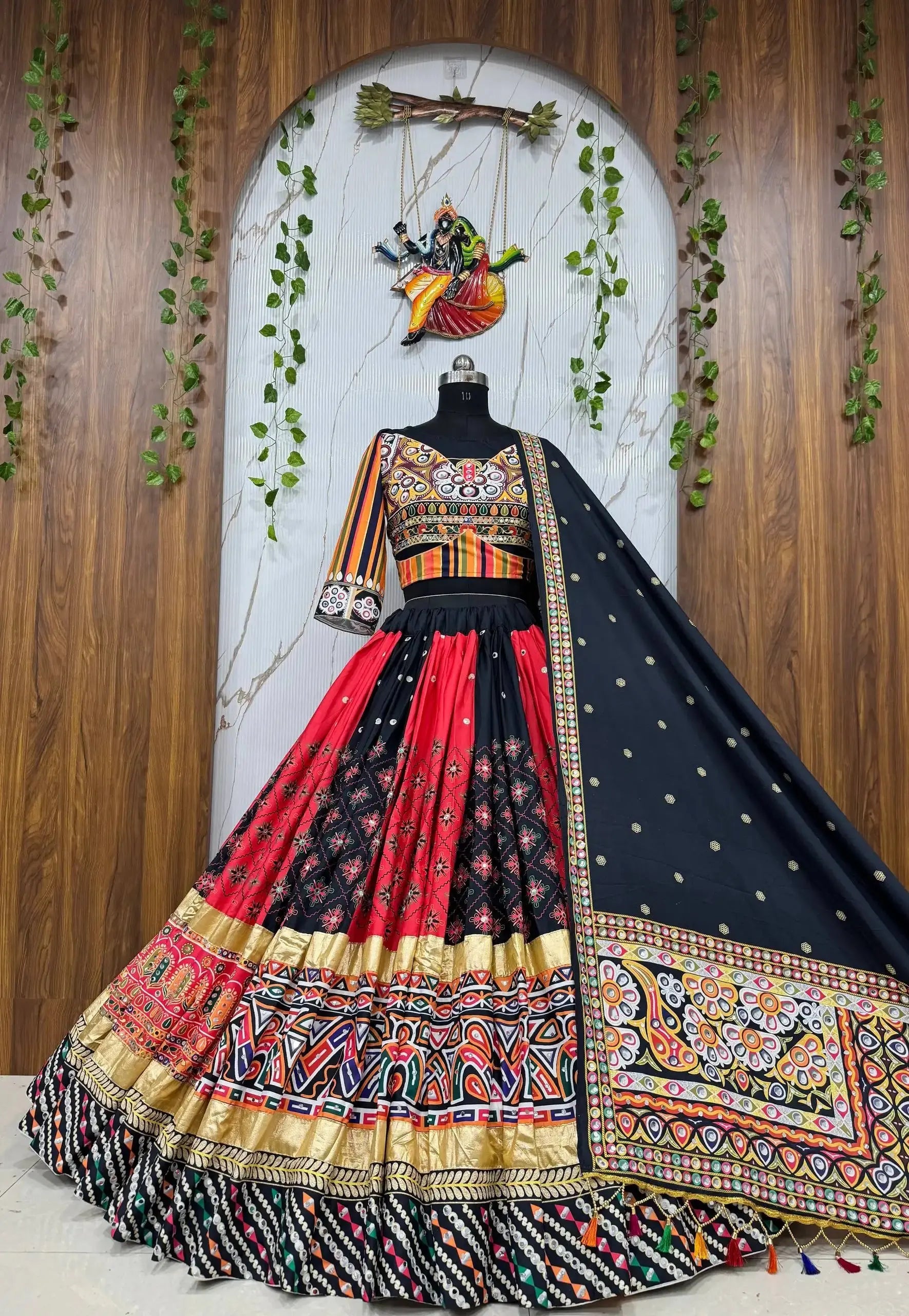 A traditional Indian lehenga choli dress displayed on a mannequin against a decorative wooden and marble backdrop. The outfit features a black and red patterned skirt with ornate gold, red, and multicolor borders, a black embroidered choli, and a black dupatta with gold motifs.