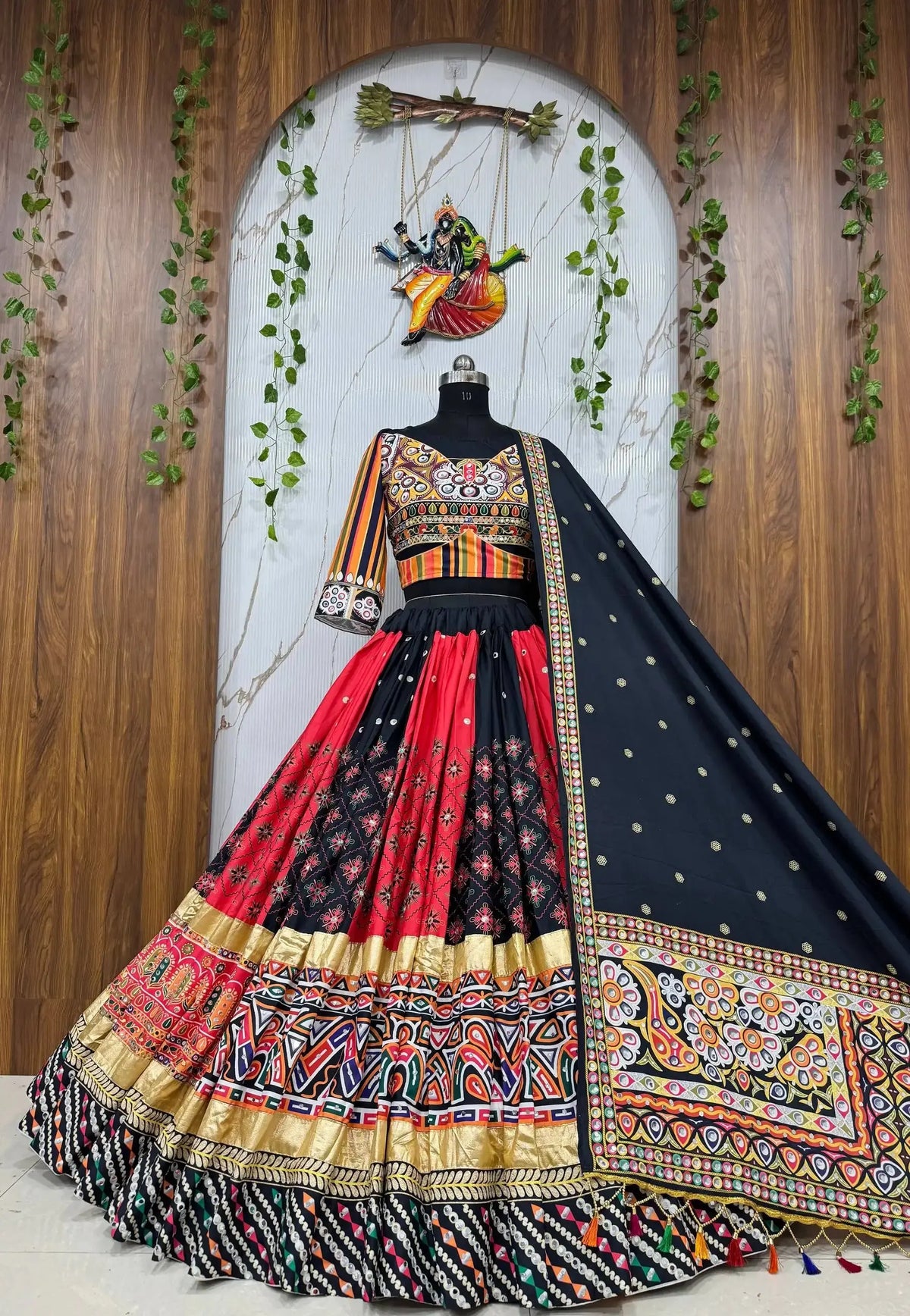A traditional Indian lehenga choli dress displayed on a mannequin against a decorative wooden and marble backdrop. The outfit features a black and red patterned skirt with ornate gold, red, and multicolor borders, a black embroidered choli, and a black dupatta with gold motifs.
