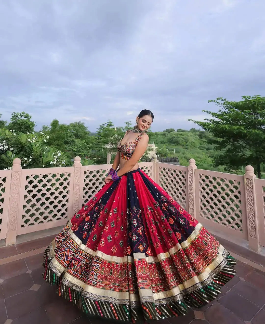 A woman wearing a voluminous red and navy lehenga choli with intricate gold and multicolor embroidery, posing outdoors on a stone terrace with a green garden background.