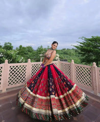 A woman wearing a voluminous red and navy lehenga choli with intricate gold and multicolor embroidery, posing outdoors on a stone terrace with a green garden background.