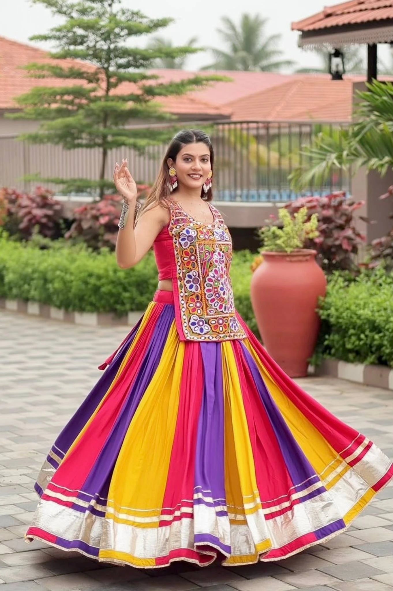 Woman in a colorful traditional outfit standing outdoors with plants and a building in the background