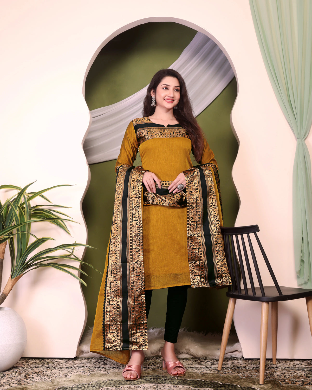 Woman wearing a mustard yellow and black traditional outfit standing in a decorative setting with plants and a chair.