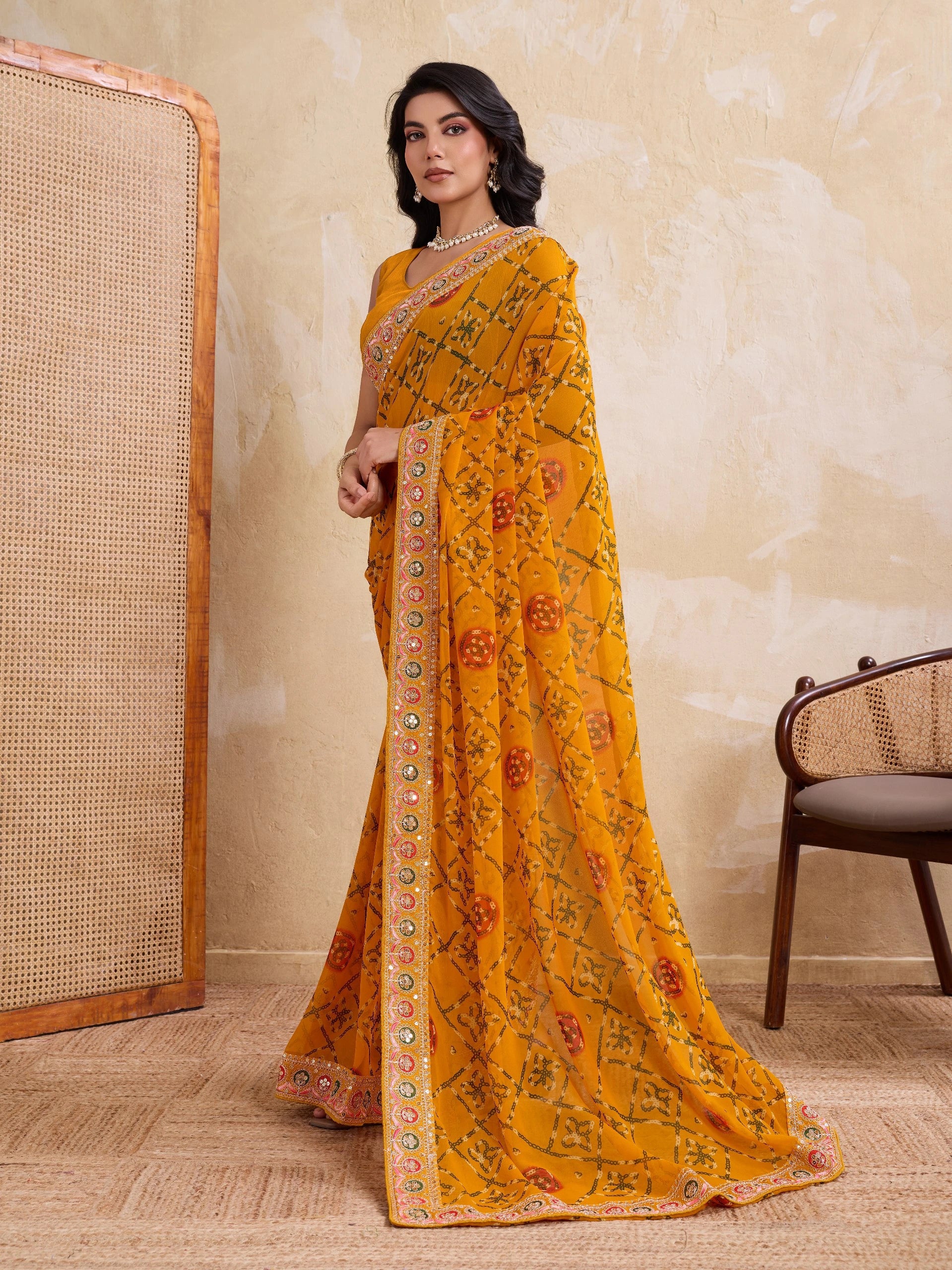 Woman wearing a yellow saree with floral patterns in a room with a textured wall and chair.