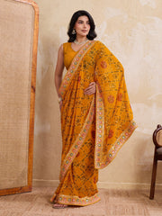 Woman wearing a yellow saree with a floral pattern against a beige wall.