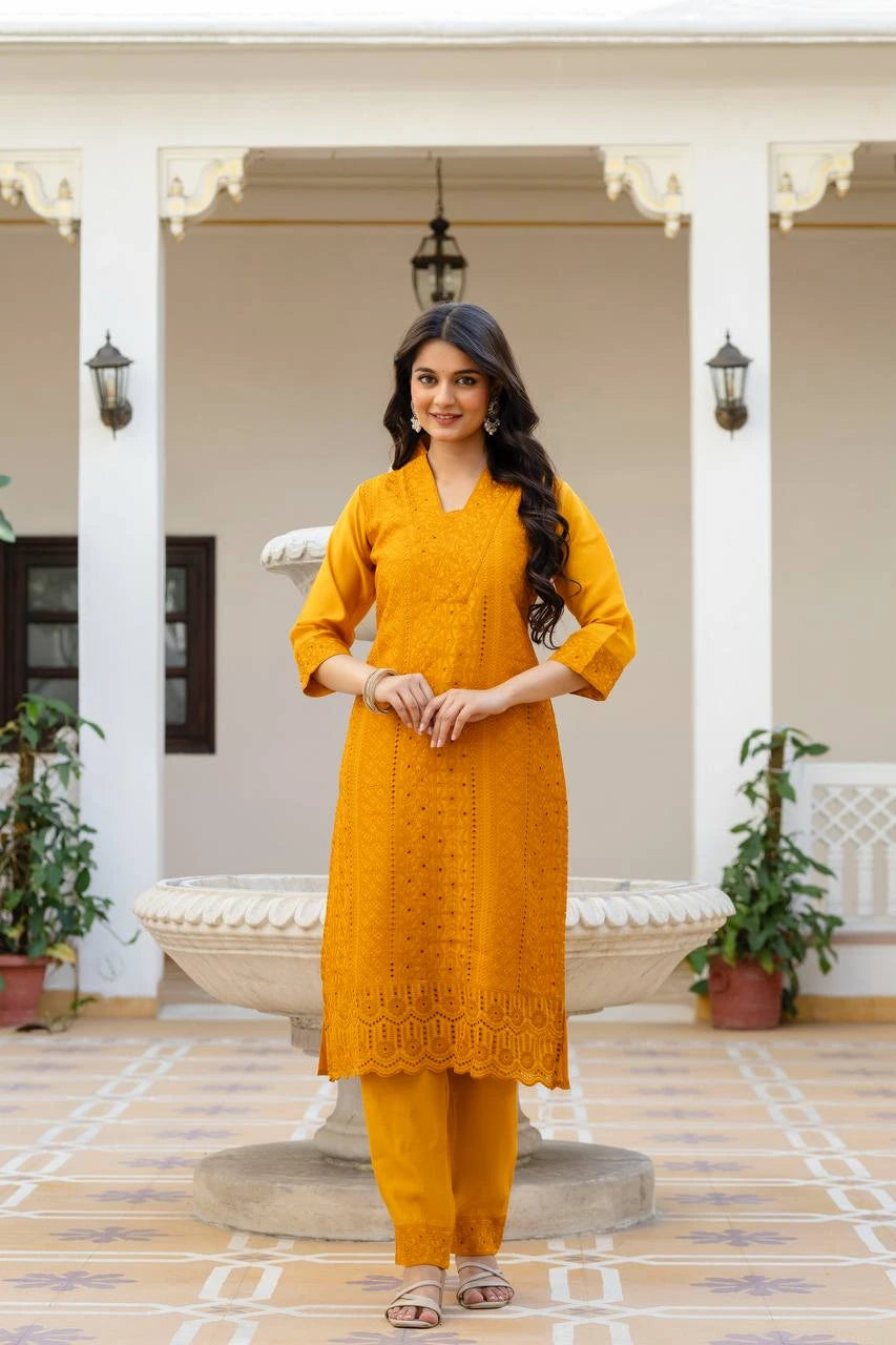 Woman in a mustard yellow traditional outfit standing in an outdoor setting with plants and a building in the background.