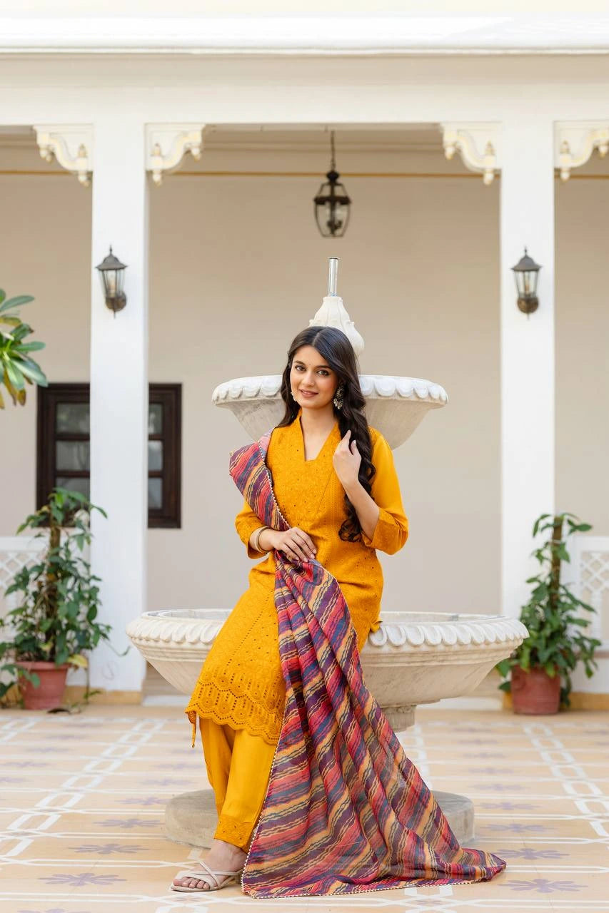 Woman in a yellow outfit holding a colorful scarf on a patio with plants and a white archway.