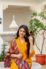 Woman in a yellow outfit with a colorful scarf standing in an indoor setting with plants.