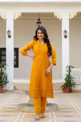 Woman in a mustard yellow traditional outfit standing in an outdoor setting with plants and a bench.