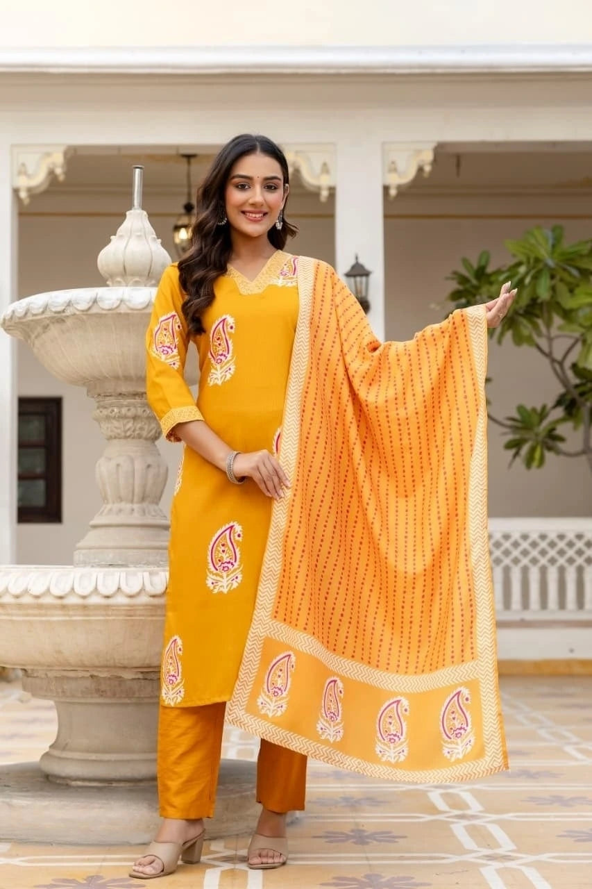 Woman in a yellow traditional outfit with an orange dupatta, standing in a decorative outdoor setting.