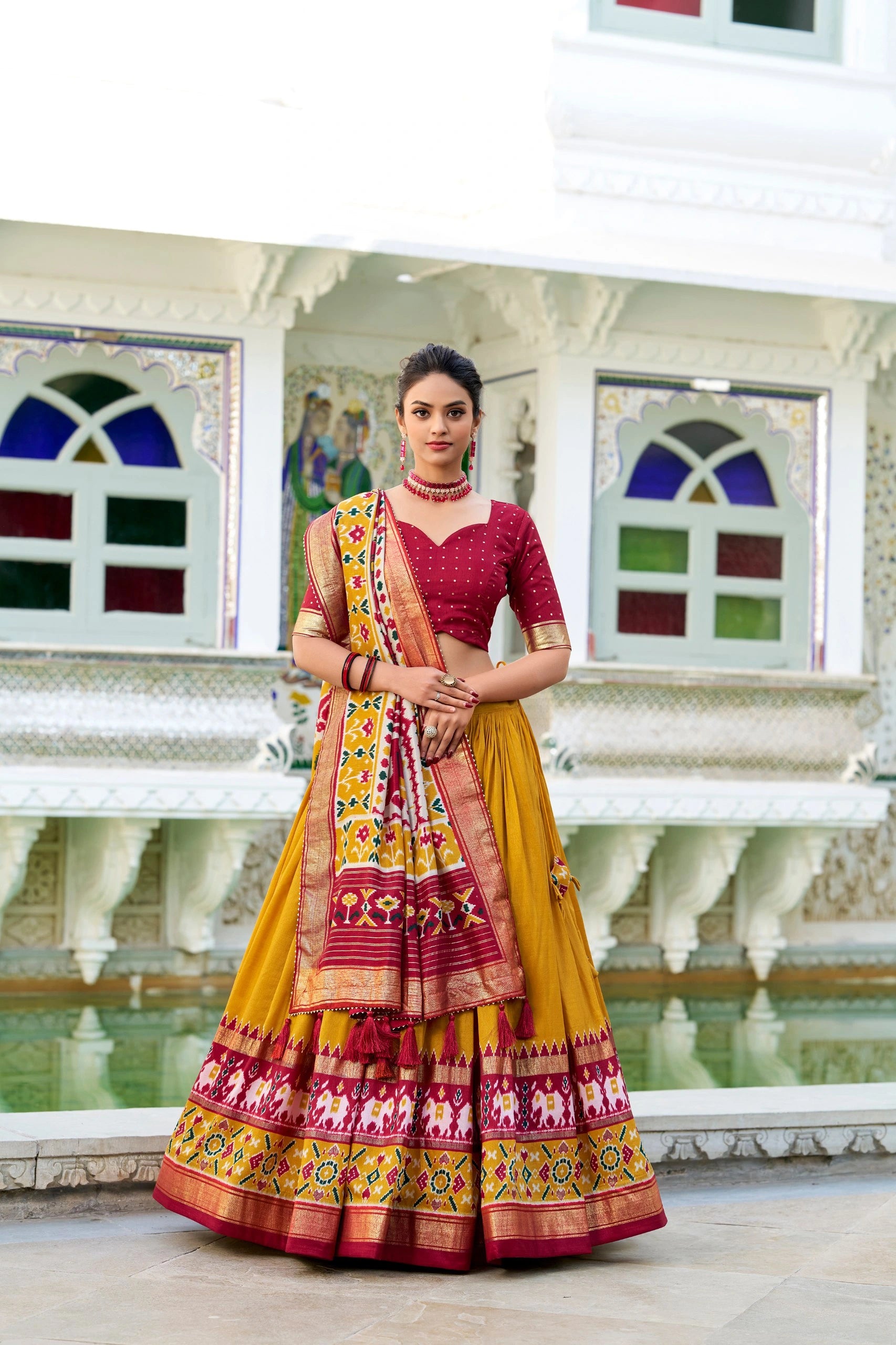 Woman in a traditional yellow and red saree with elephant patterns, standing in front of a decorative wall.