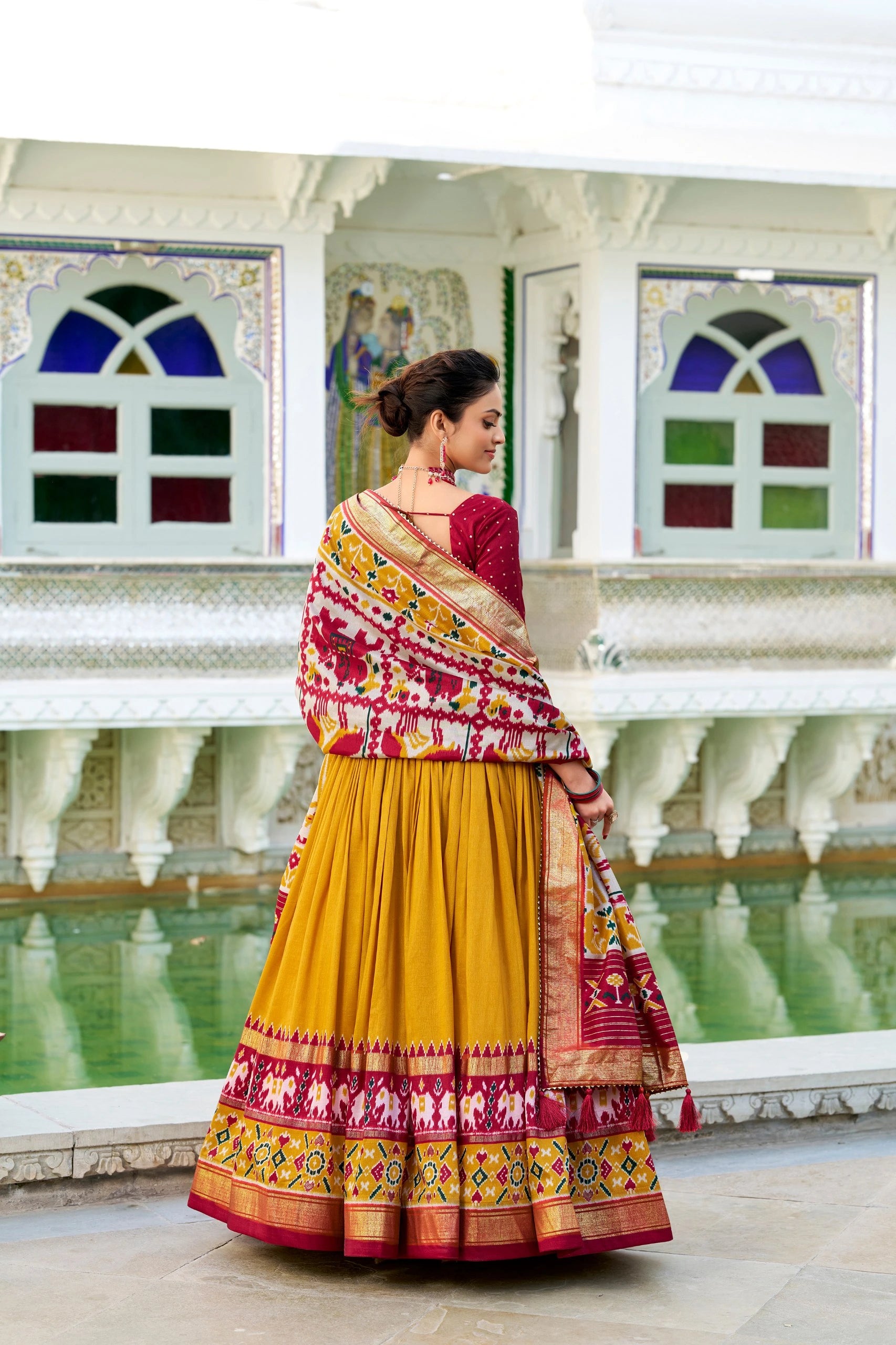 Woman in a traditional yellow and multicolored saree standing in front of a decorative wall with stained glass windows.
