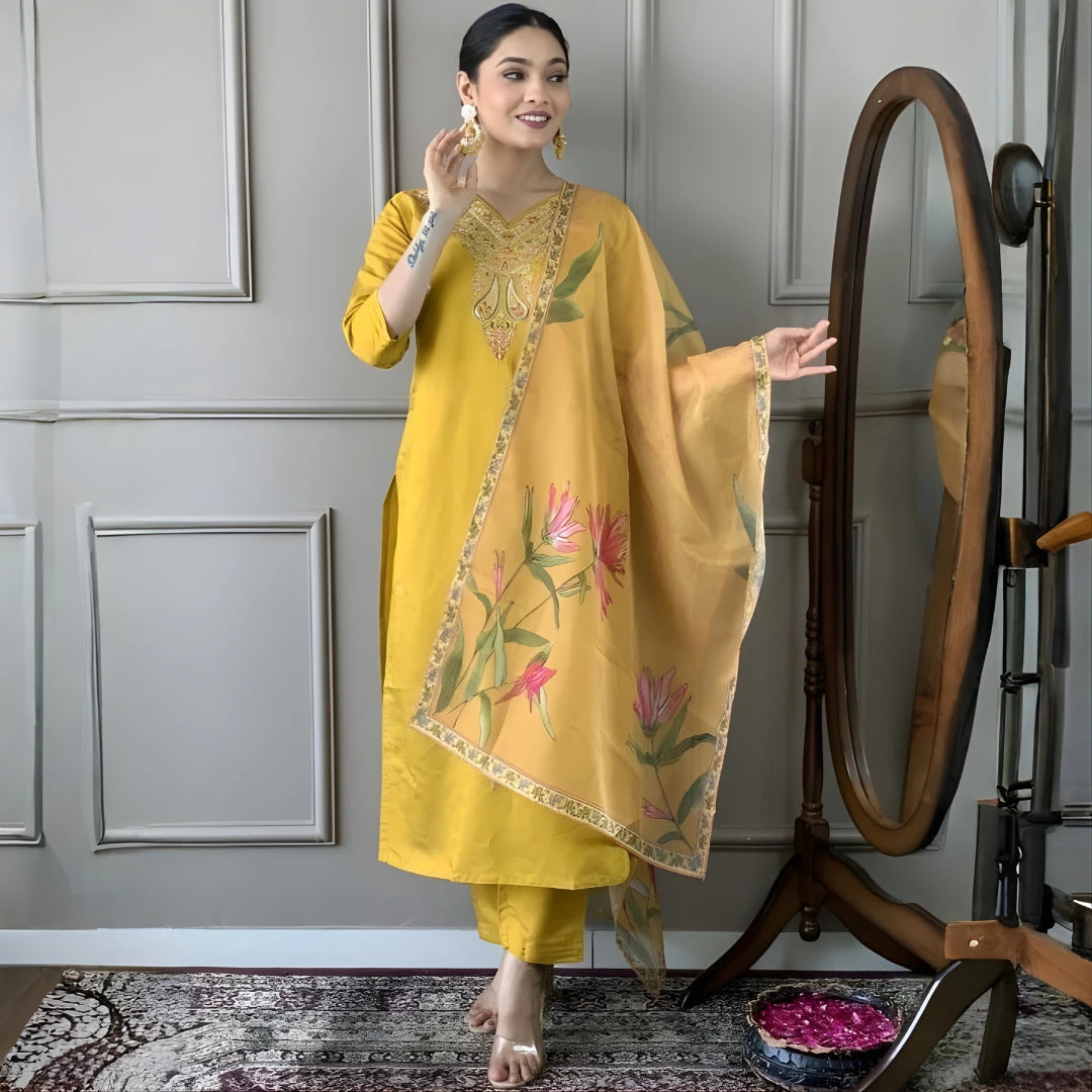 Woman in a yellow traditional outfit with floral design standing next to a wooden mirror against a gray wall.