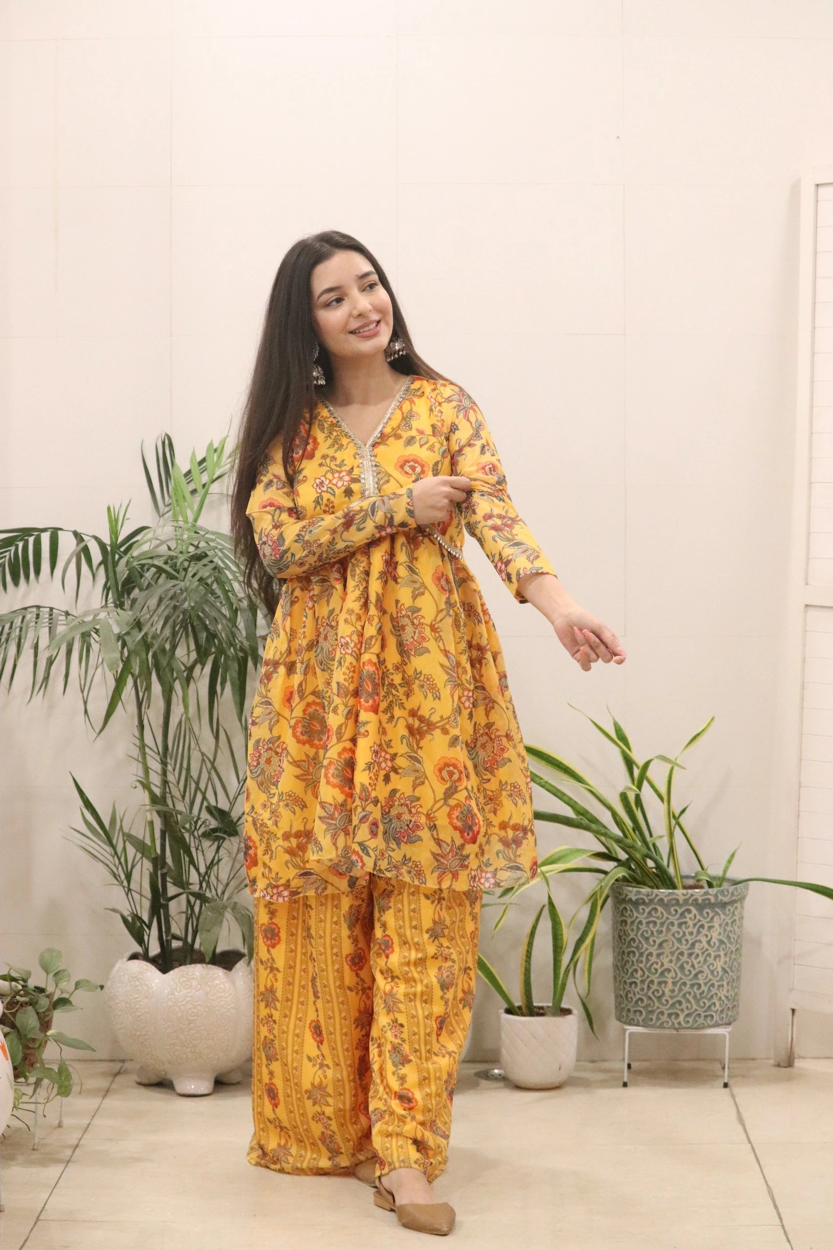 Woman wearing a yellow floral outfit standing indoors with plants in the background